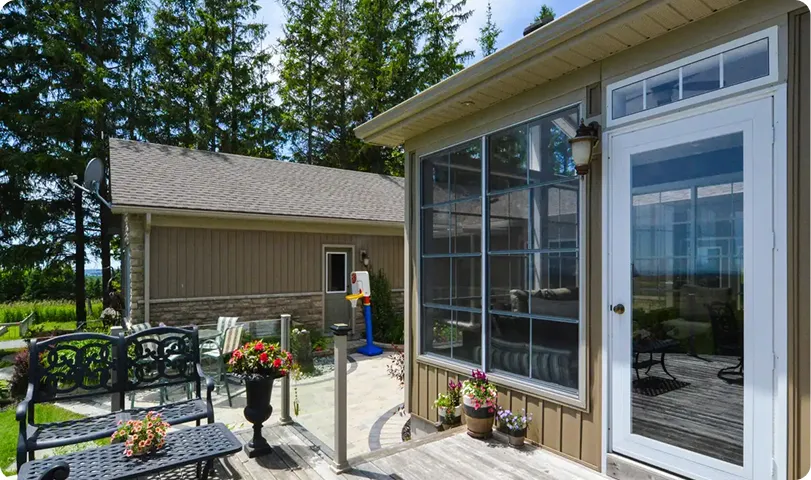 Outdoor patio area with a metal bench, a small table with flowers, potted plants, and a large windowed door of a beige house.