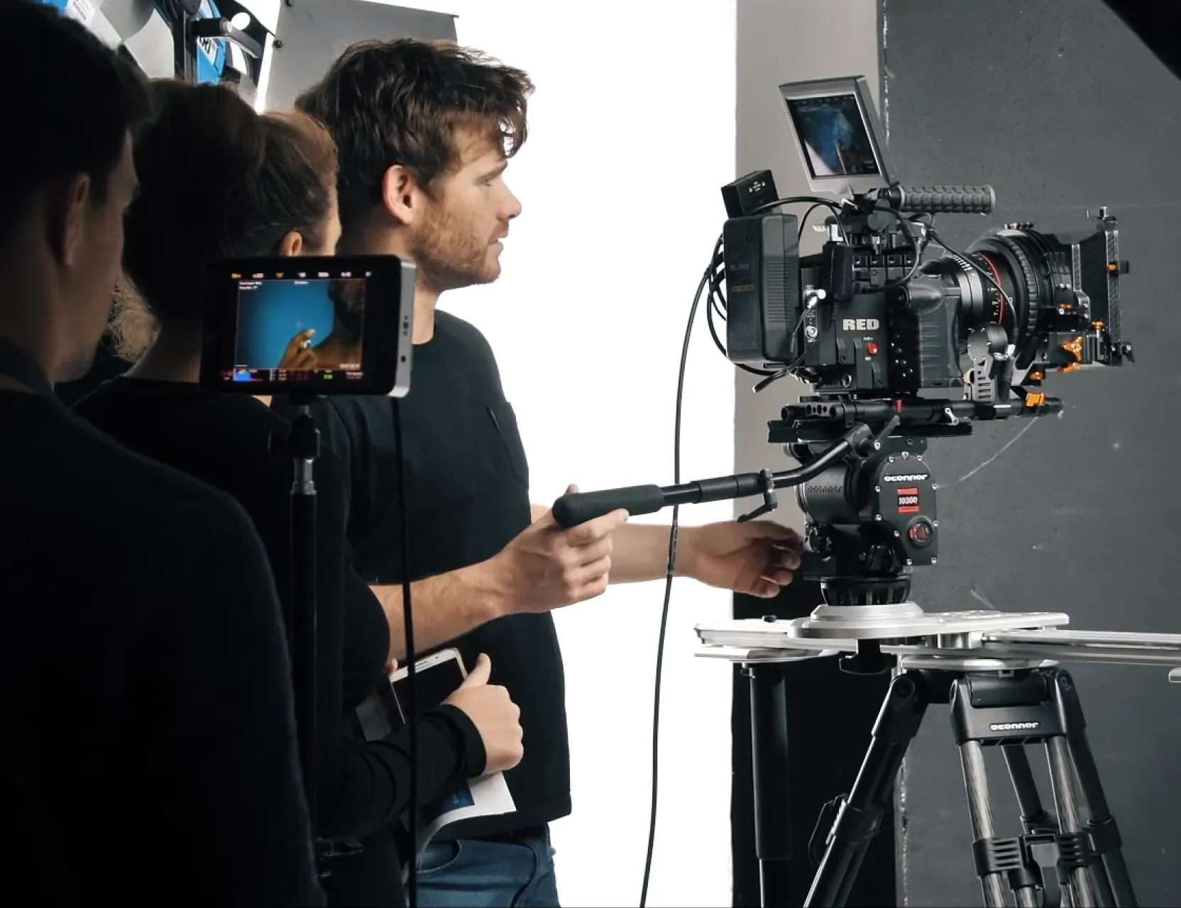 Camera operator adjusts a RED cinema camera on a tripod in a white studio as crew look on.