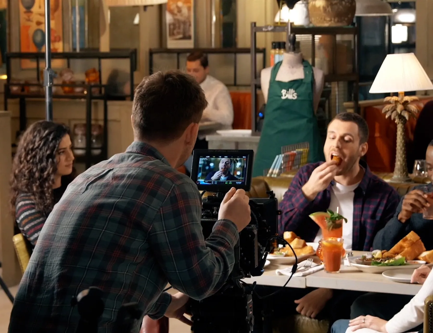 Camera operator films a group enjoying a meal together at a restaurant table, checking the shot on a small monitor.