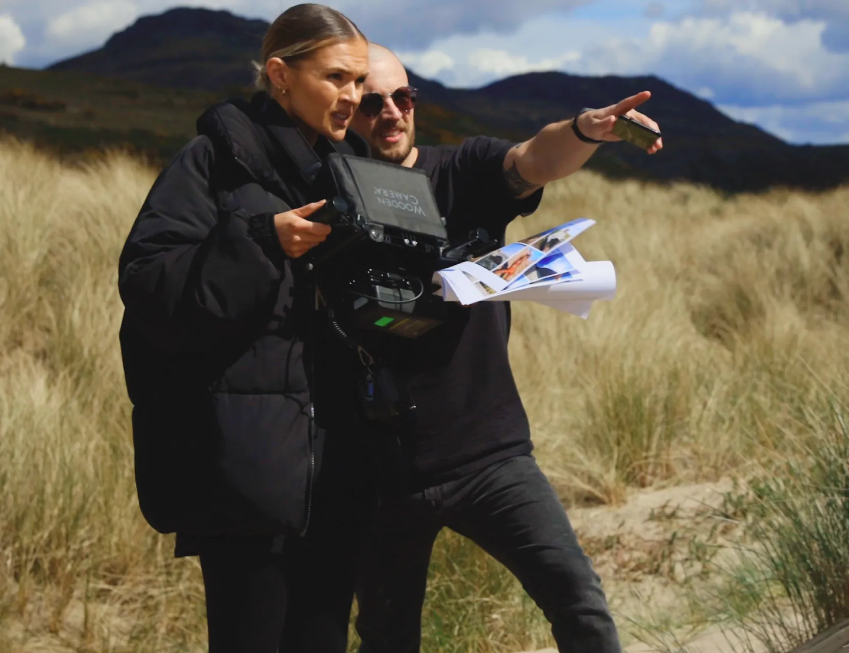 Two crew members on a grassy dune with mountains behind them, one operating a handheld camera gimbal while the other points off-frame.