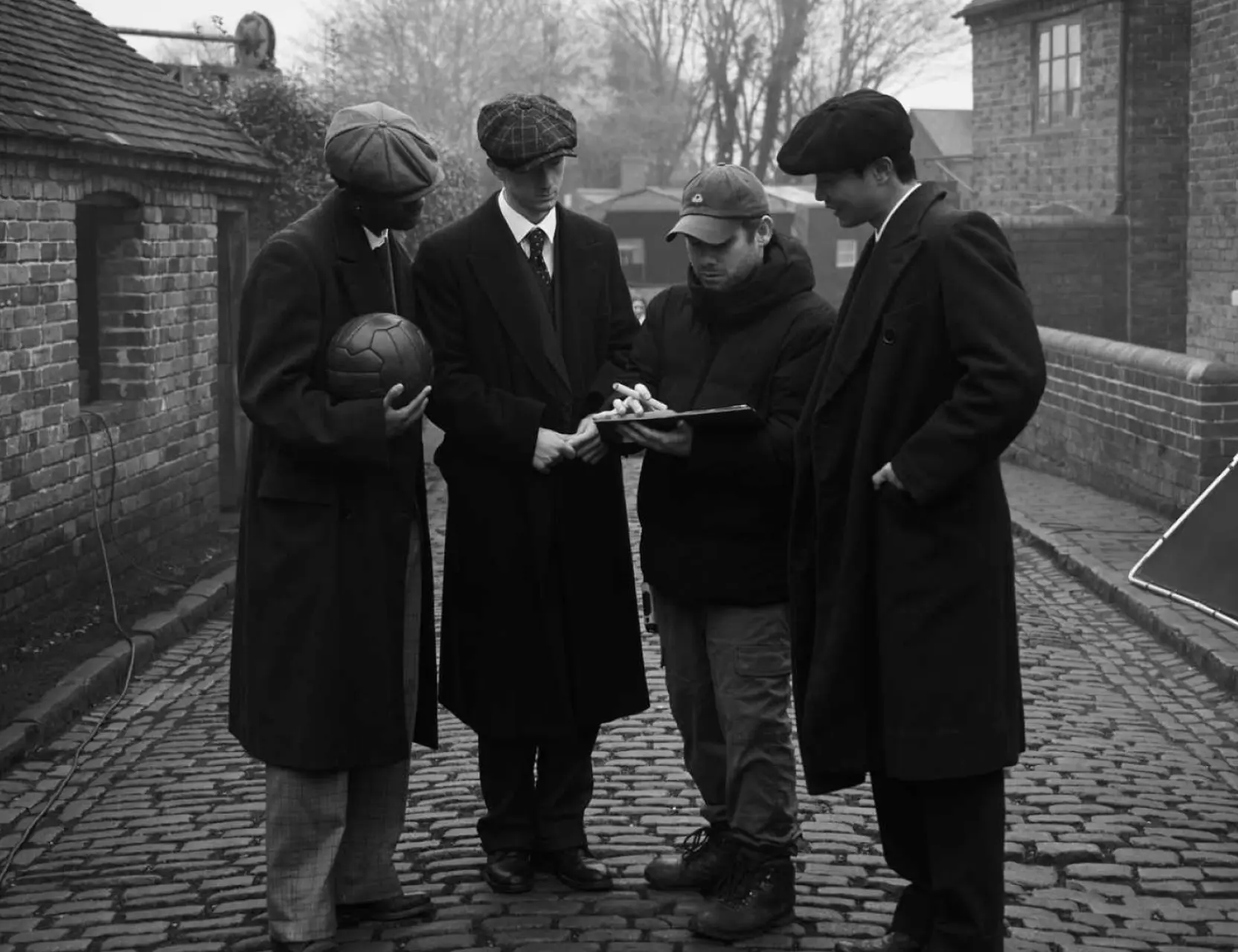 Four men in vintage flat caps and long coats stand on a cobbled street during a 1920s-style period shoot, one crew member checking notes.