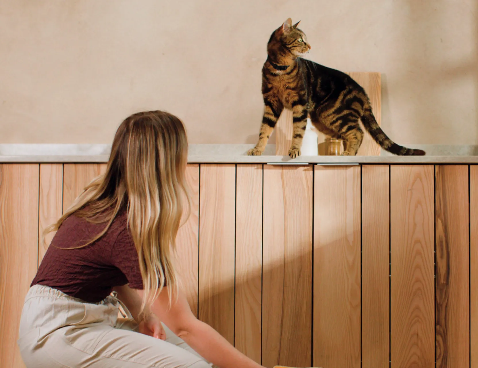 Woman kneeling beside a wooden unit watches a tabby cat perched on top during a product shoot.