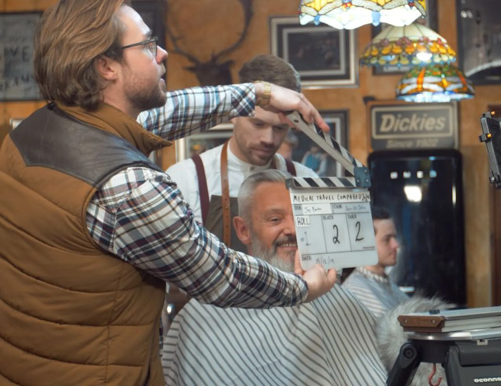 Crew member marks the clapperboard on a barbershop film set as a barber cuts a client's hair.