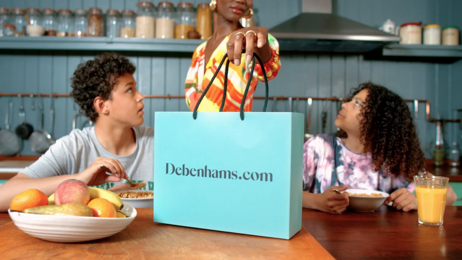 Two children sitting at a kitchen table having breakfast with cereal and juice, looking up at a woman handing them a large blue shopping bag labeled Debenhams.com.