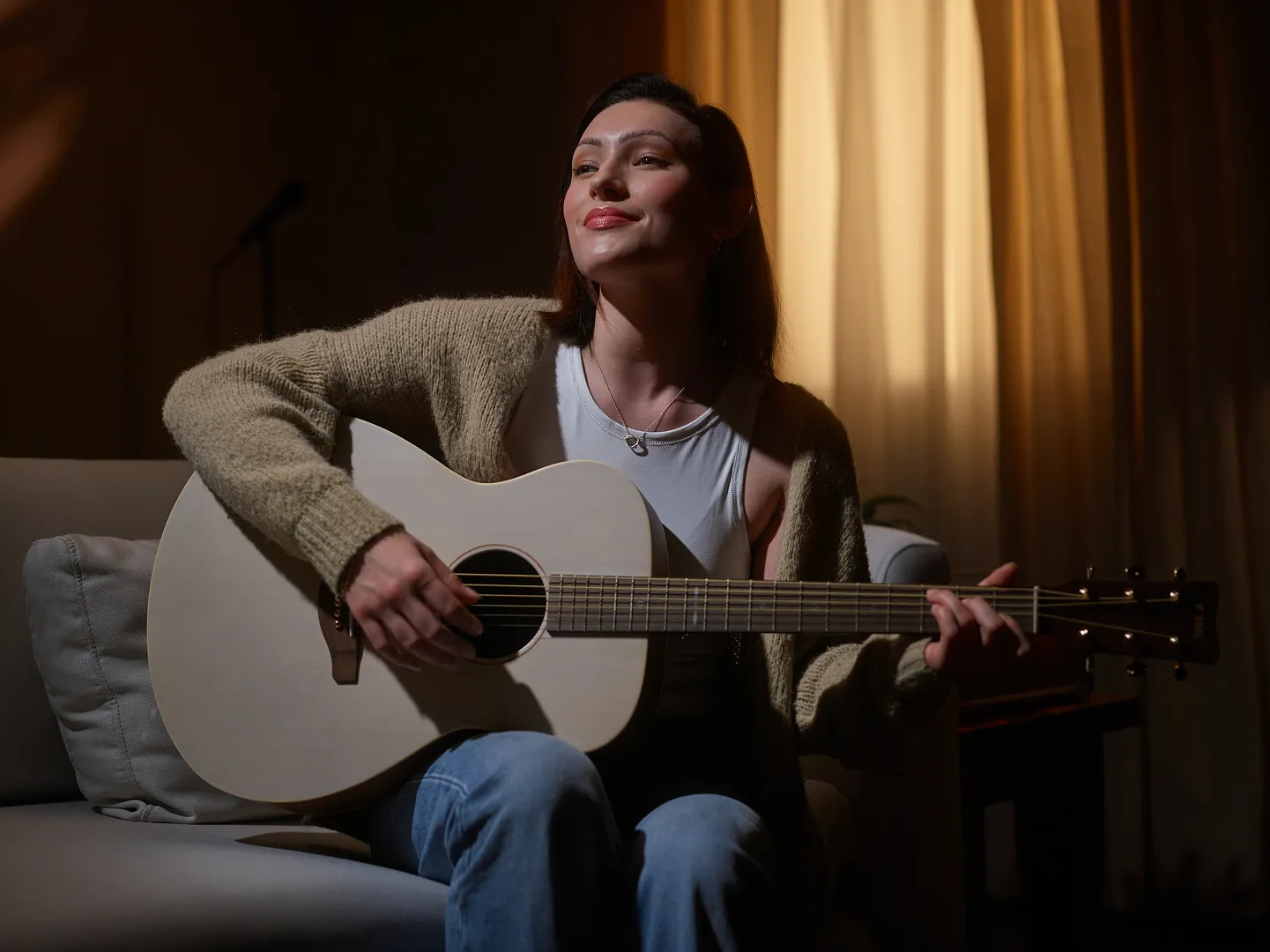 A woman sitting on a sofa playing an acoustic guitar with a calm expression, softly lit by warm indoor lighting with curtains in the background.
