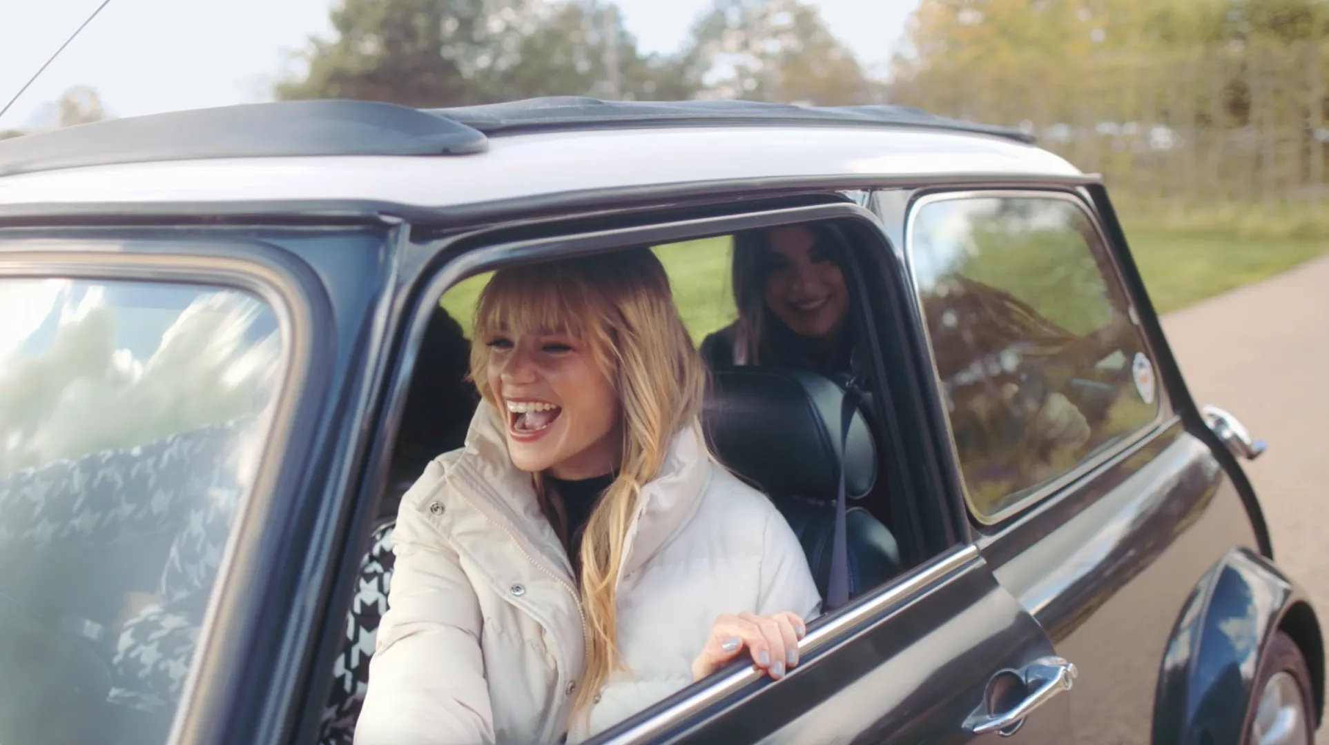 Young woman with blonde hair laughing while sitting in the open window of a dark-colored classic car on a road with greenery in the background, with another smiling woman visible inside the car.