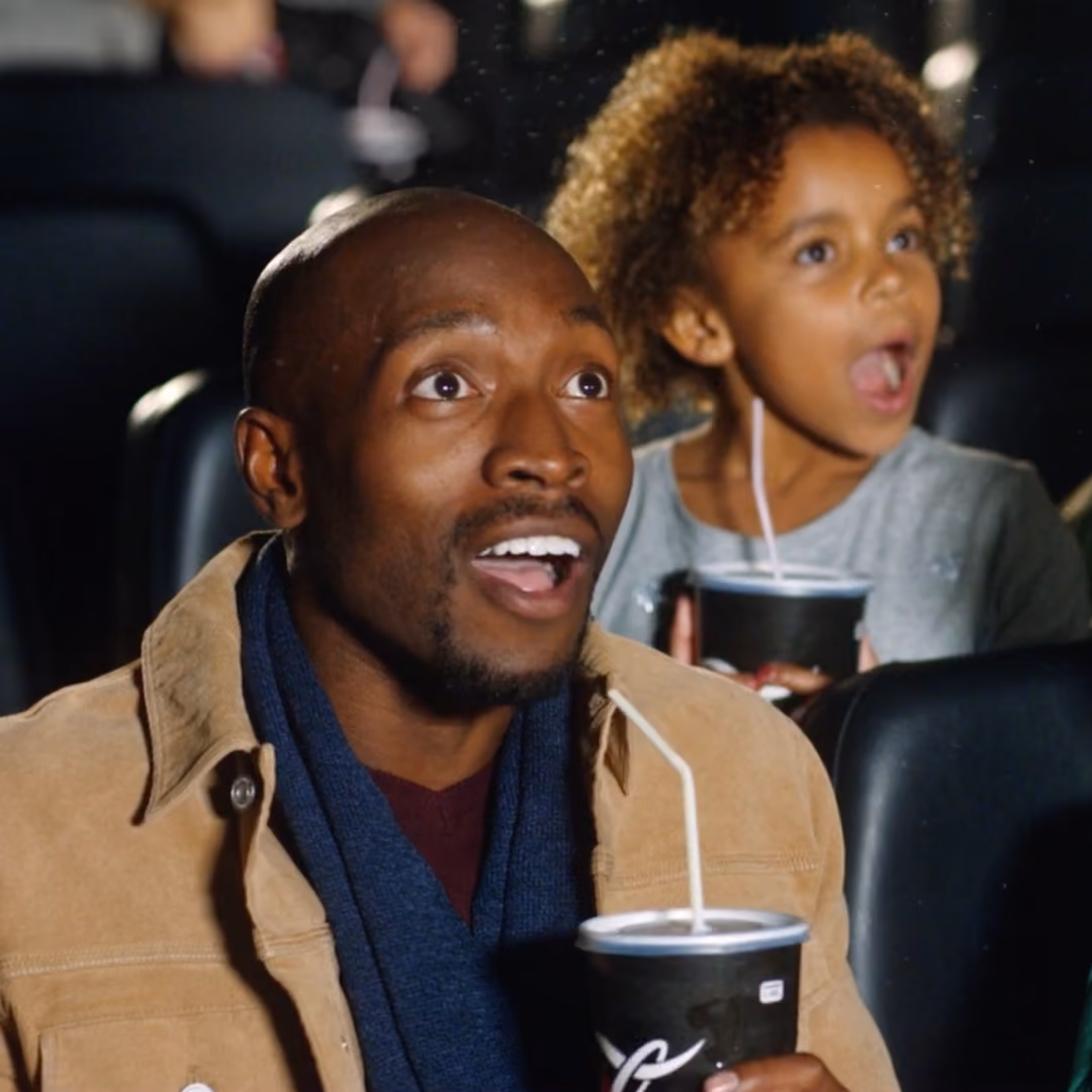 Father and daughter watching wide-eyed in cinema seats, each holding a drink with a straw.