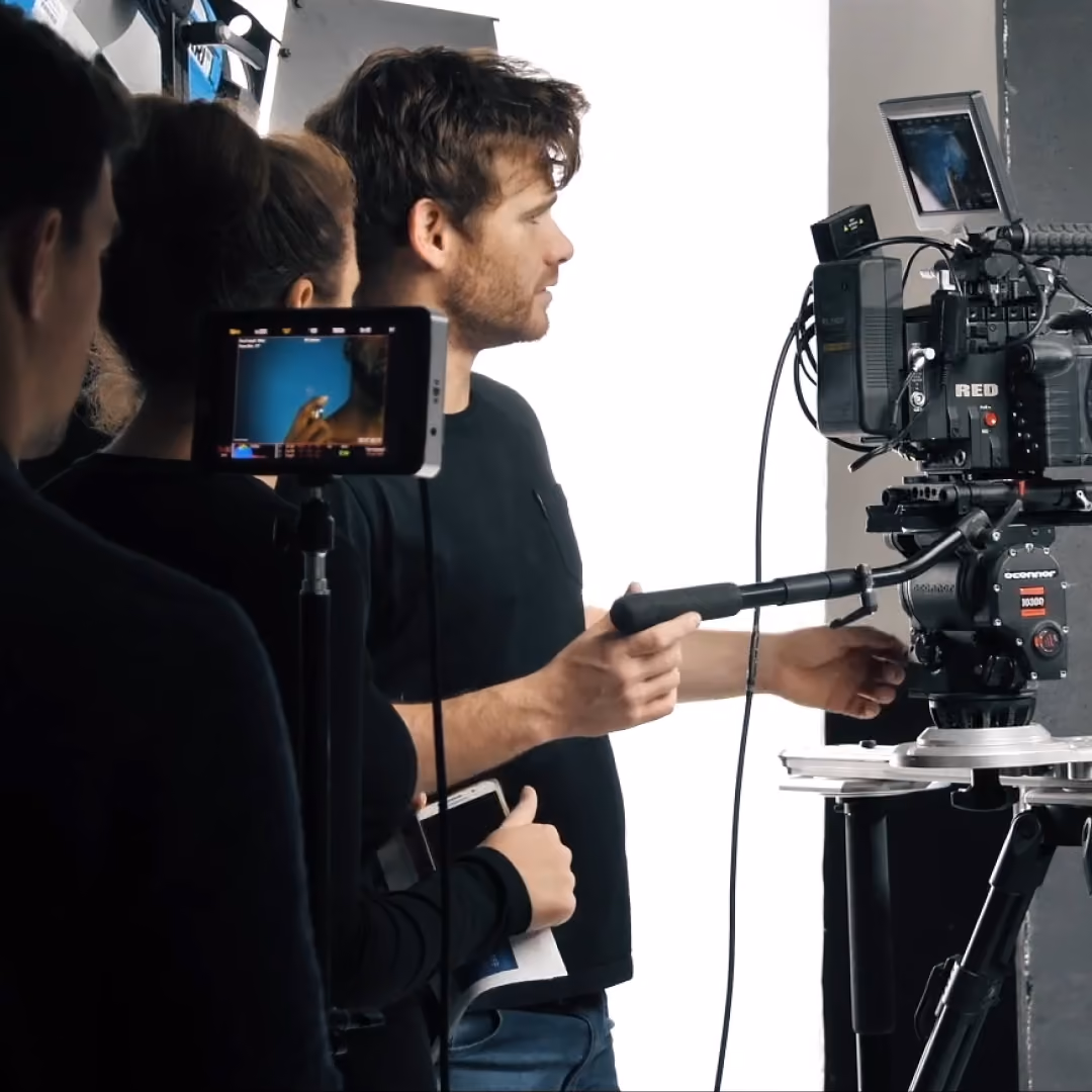 Film crew on set operating a RED cinema camera and monitor as a man is filmed against a plain studio backdrop.