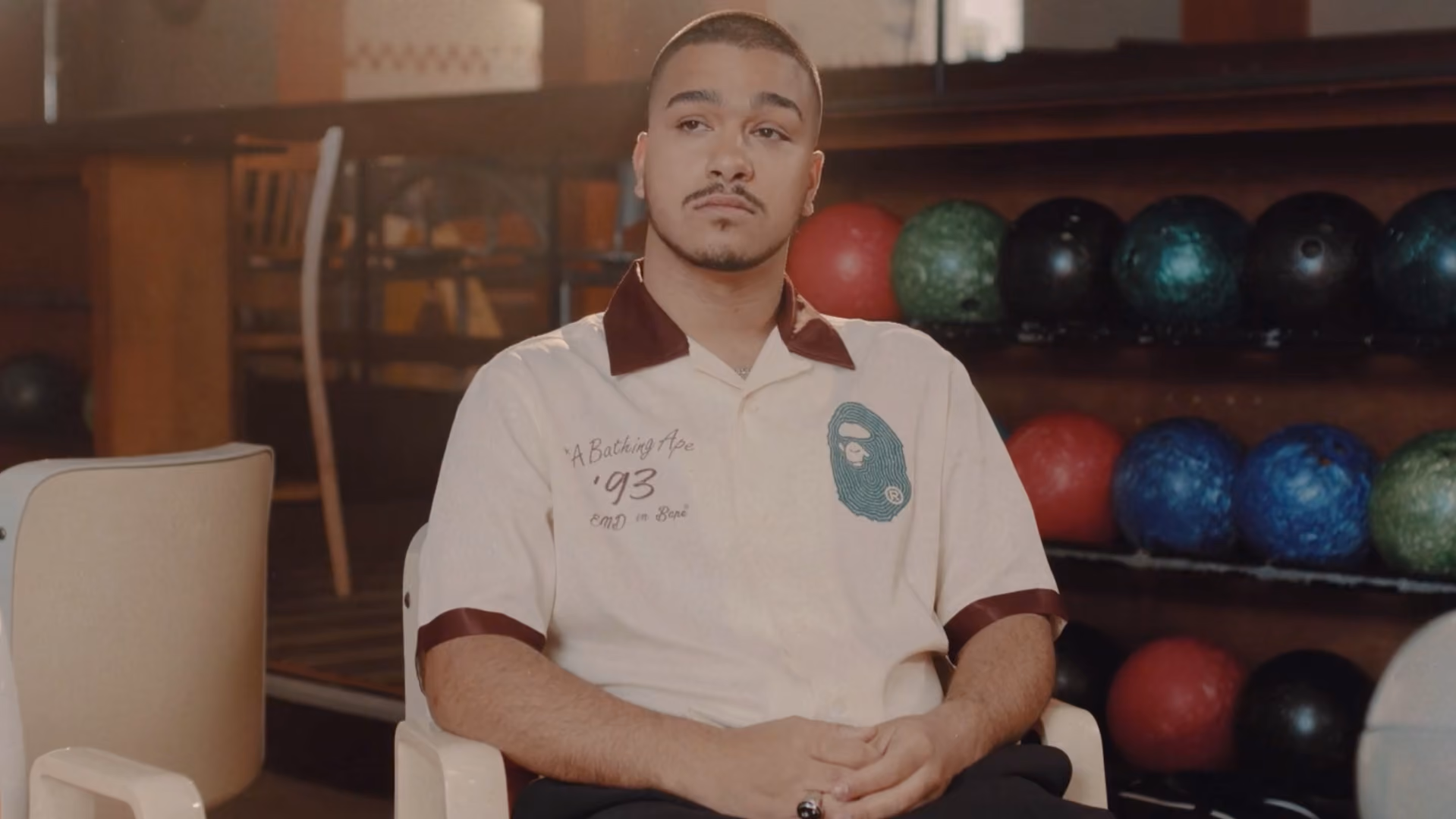 A man with short hair and a mustache sits with hands clasped, wearing a white shirt with a green graphic on chest, in front of a rack of colorful bowling balls inside a bowling alley.