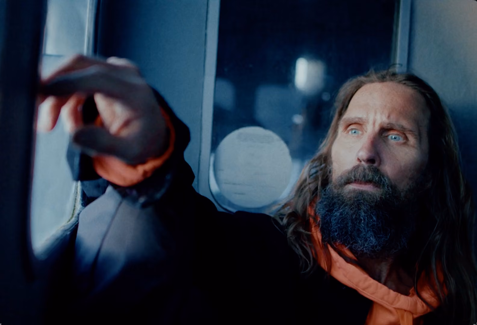 Man with long hair and a beard wearing a dark jacket with an orange hood, looking intently outside a window while reaching towards it.