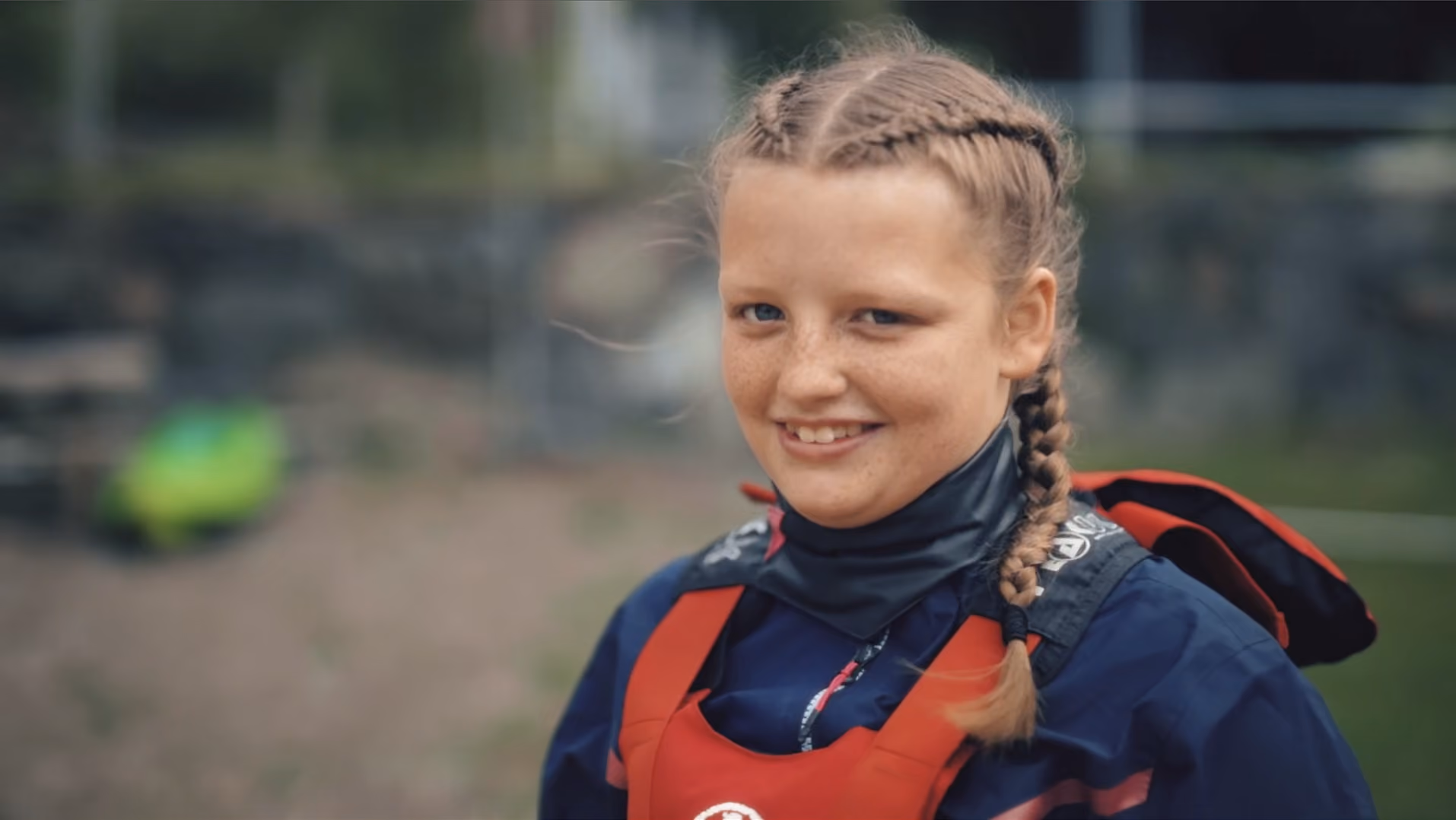 Young girl with braided hair wearing a dark jacket and a red life vest, smiling outdoors with a blurred background.
