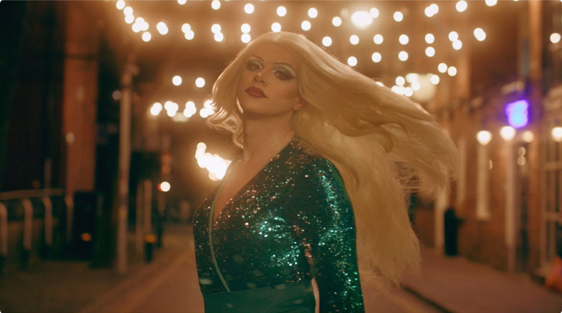 Person with long blonde hair wearing a shiny green sequined outfit, posing with head turned slightly and street lights glowing softly in the background.