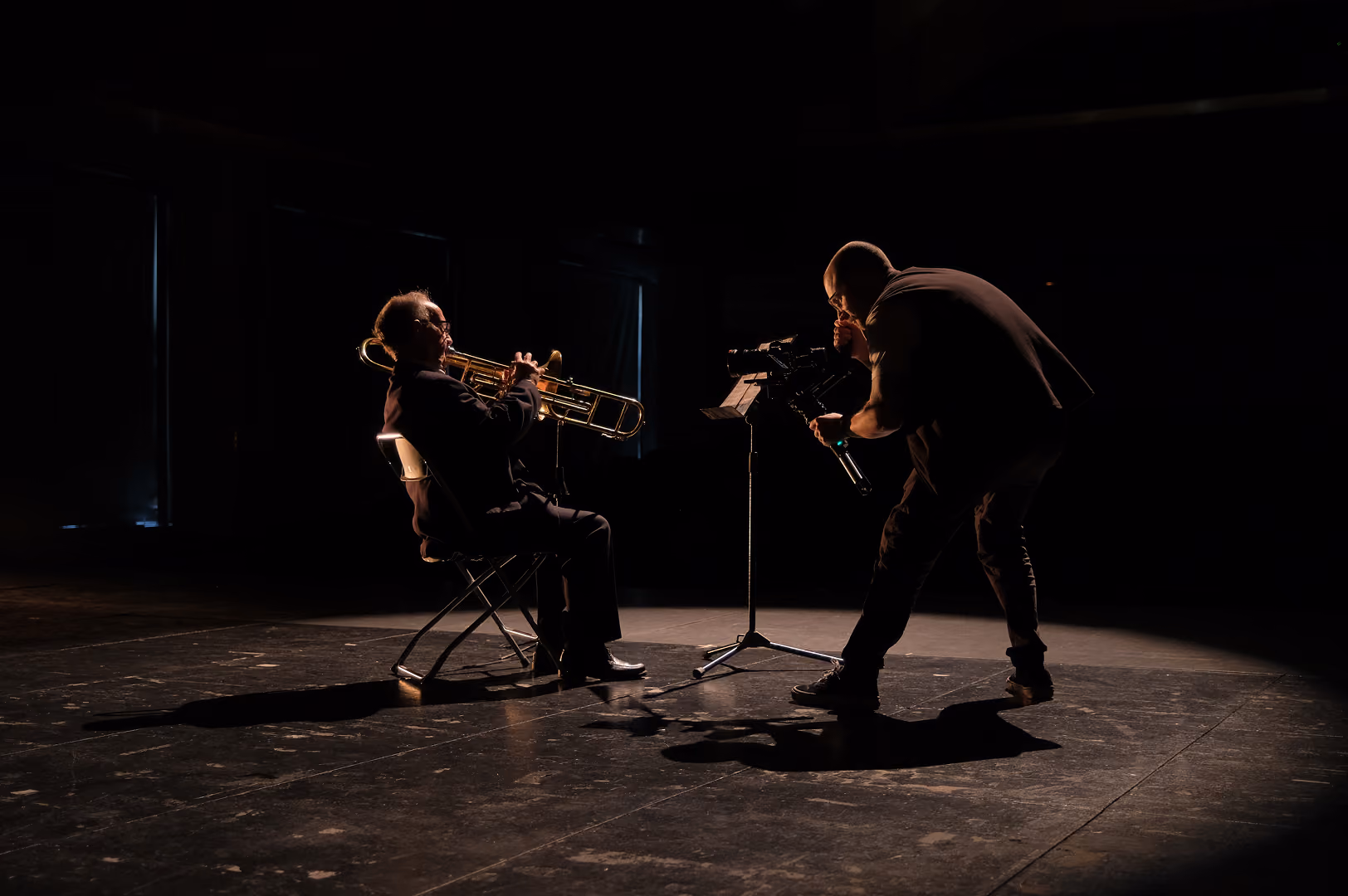 A seated musician playing a trombone under a spotlight on a dark stage while a cameraman films closely with a professional camera.