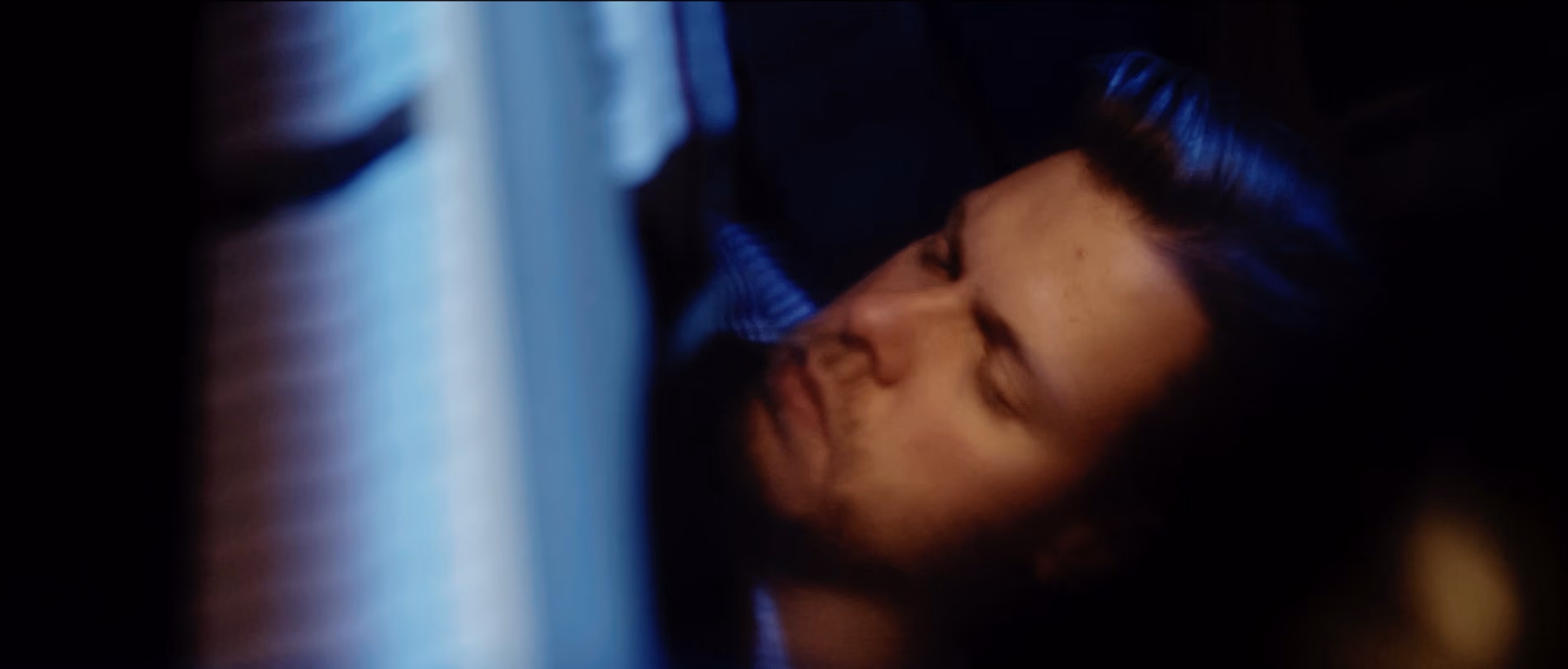 Close-up of a man with closed eyes illuminated by soft blue light, his face partially obscured by an out-of-focus object in the foreground.