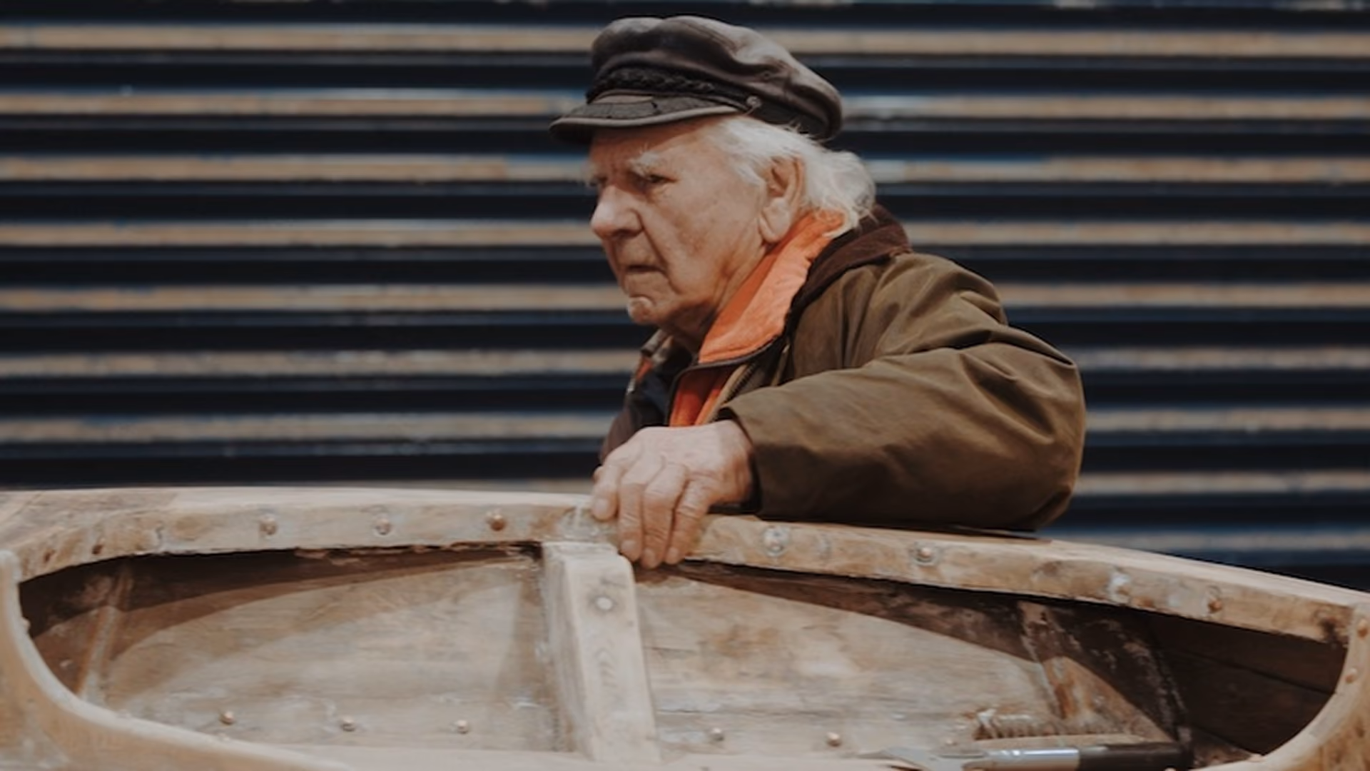Elderly man with white hair wearing a dark cap and brown jacket holding the side of a rustic wooden boat in front of a dark horizontally-striped wall.