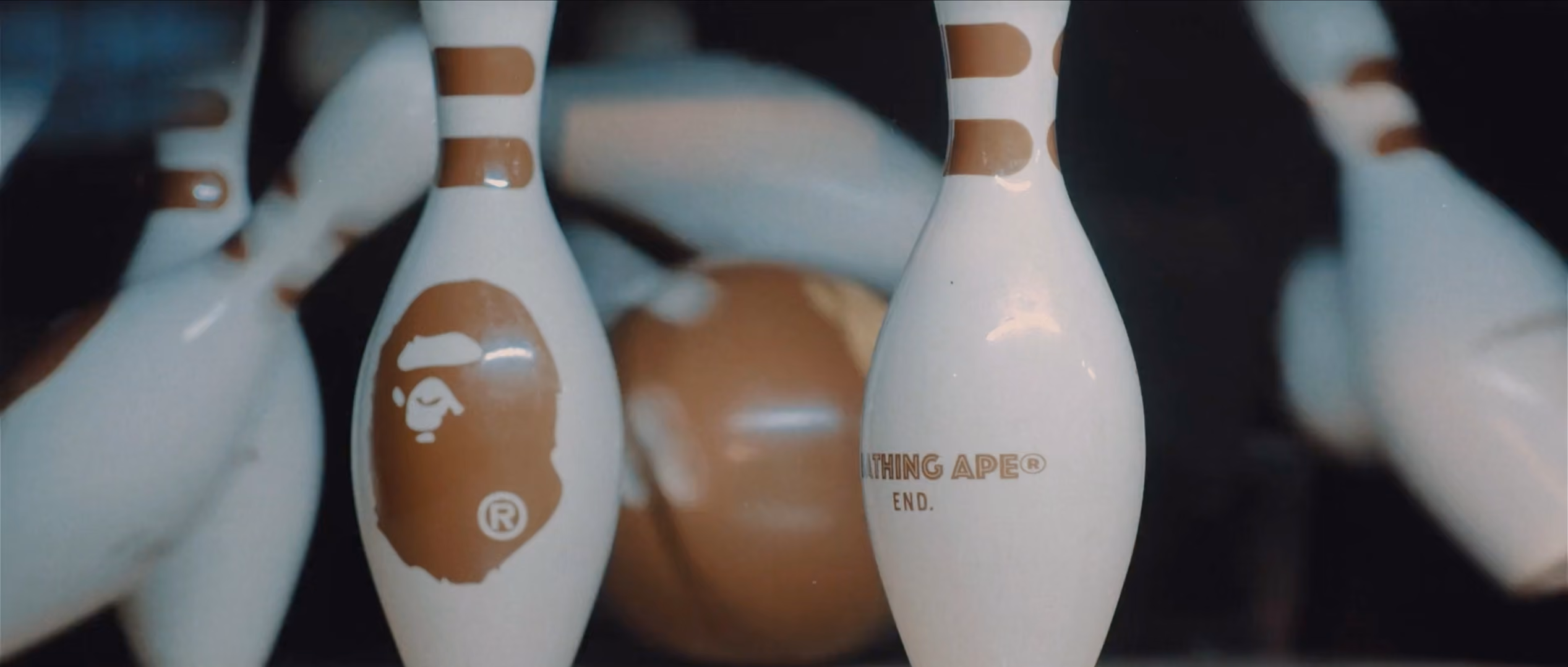 Close-up of white bowling pins with brown stripe designs, one pin features the 'A Bathing Ape' logo and the text 'A BATHING APE® END.'