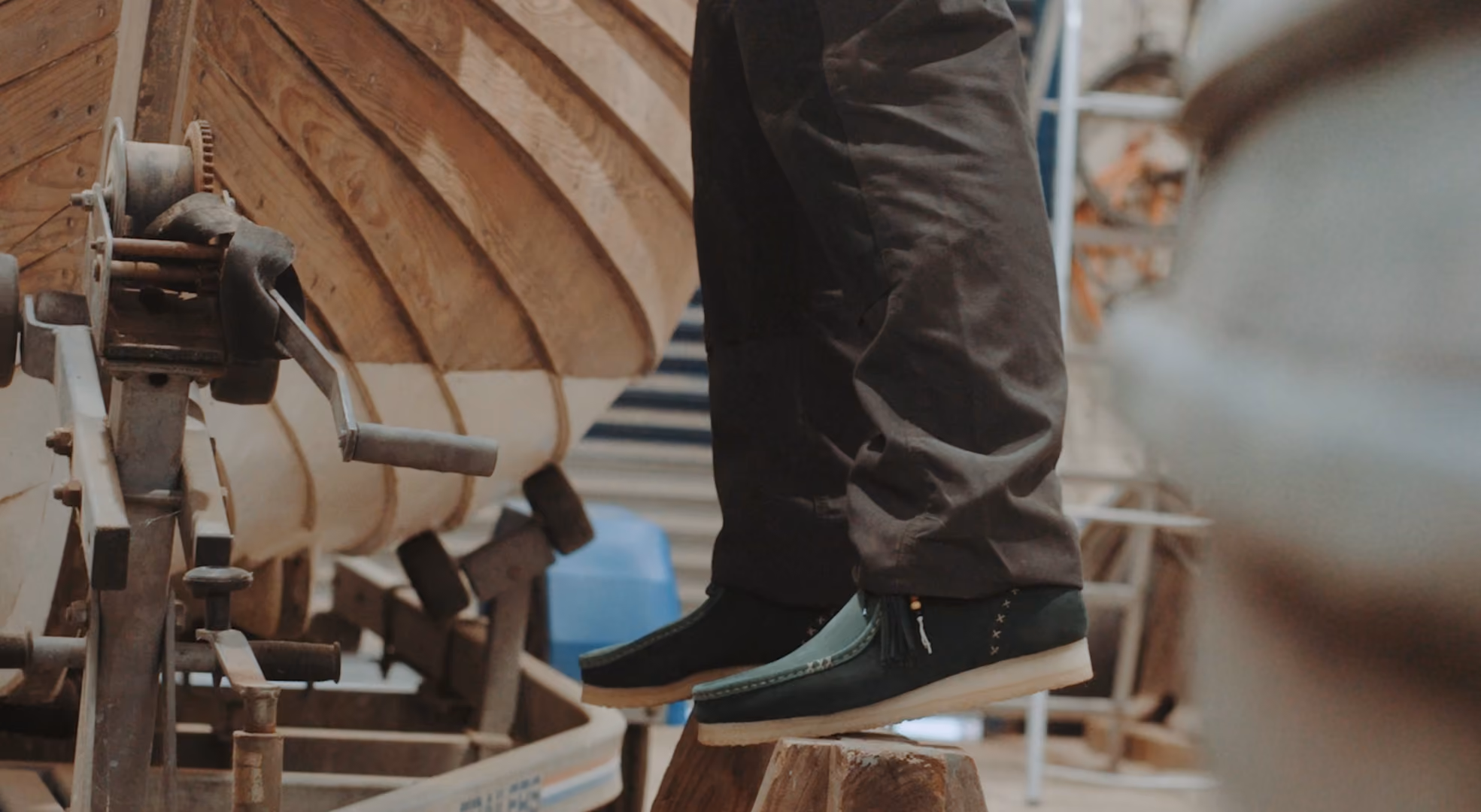 Person wearing dark pants and black slip-on shoes standing on wooden blocks in a workshop with a wooden boat hull and tools in the background.