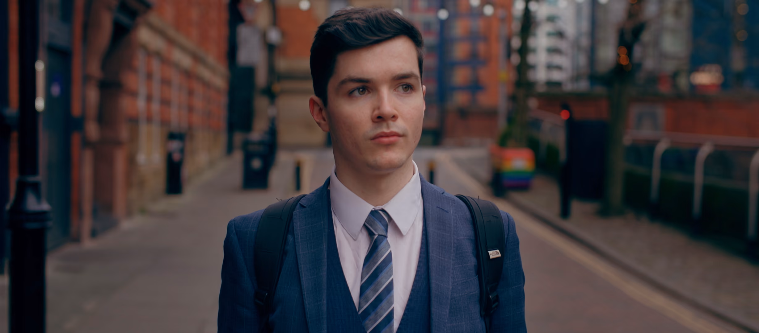 Young man in a blue suit, light blue shirt, and striped tie walking on a city street with backpack straps over his shoulders, surrounded by buildings and blurred background.