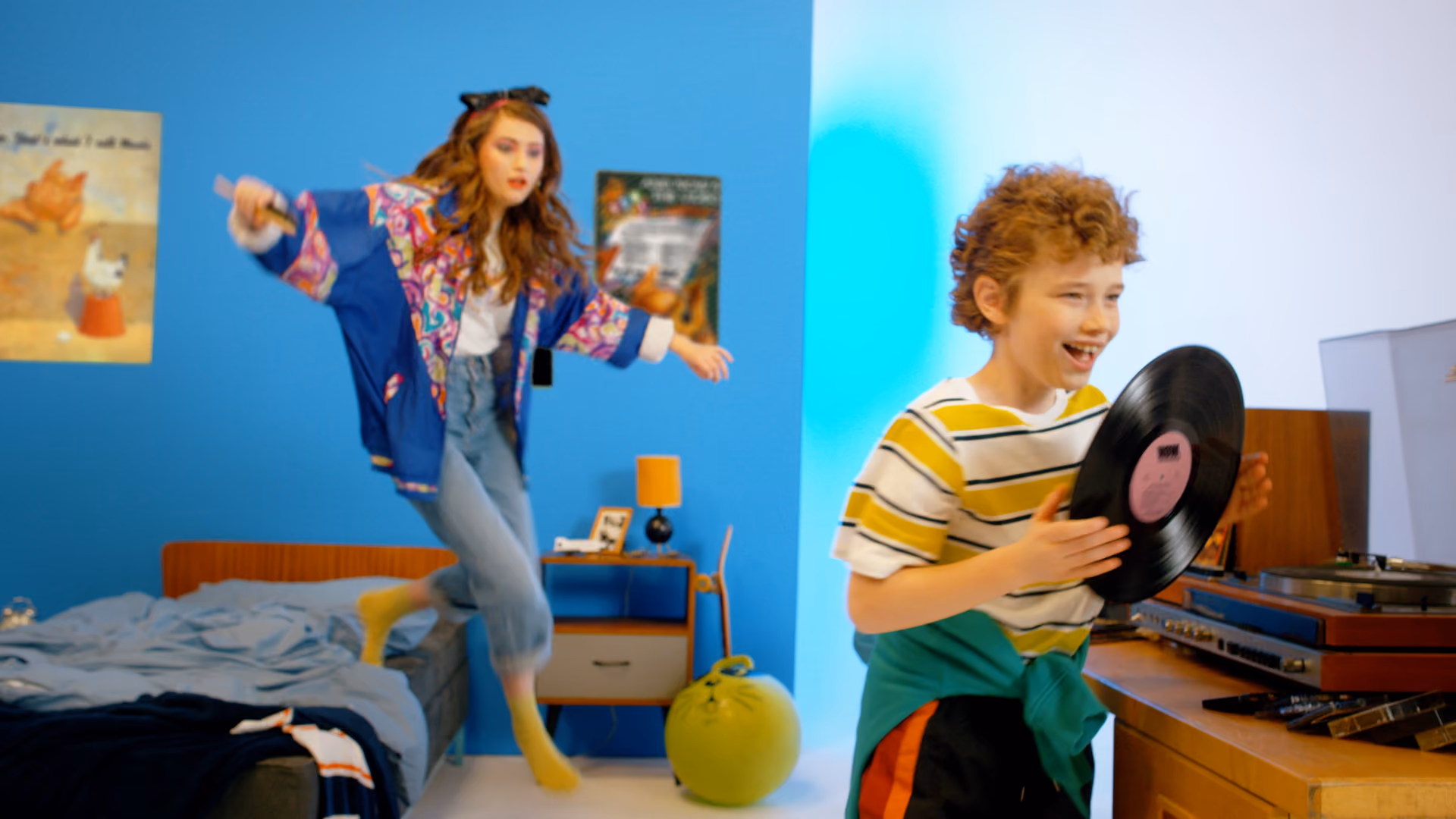 A boy with curly hair wearing a striped shirt and green sweater around his waist holds a vinyl record smiling near a turntable, while a girl with long hair in colorful clothes jumps off a bed in a room with blue and white walls.