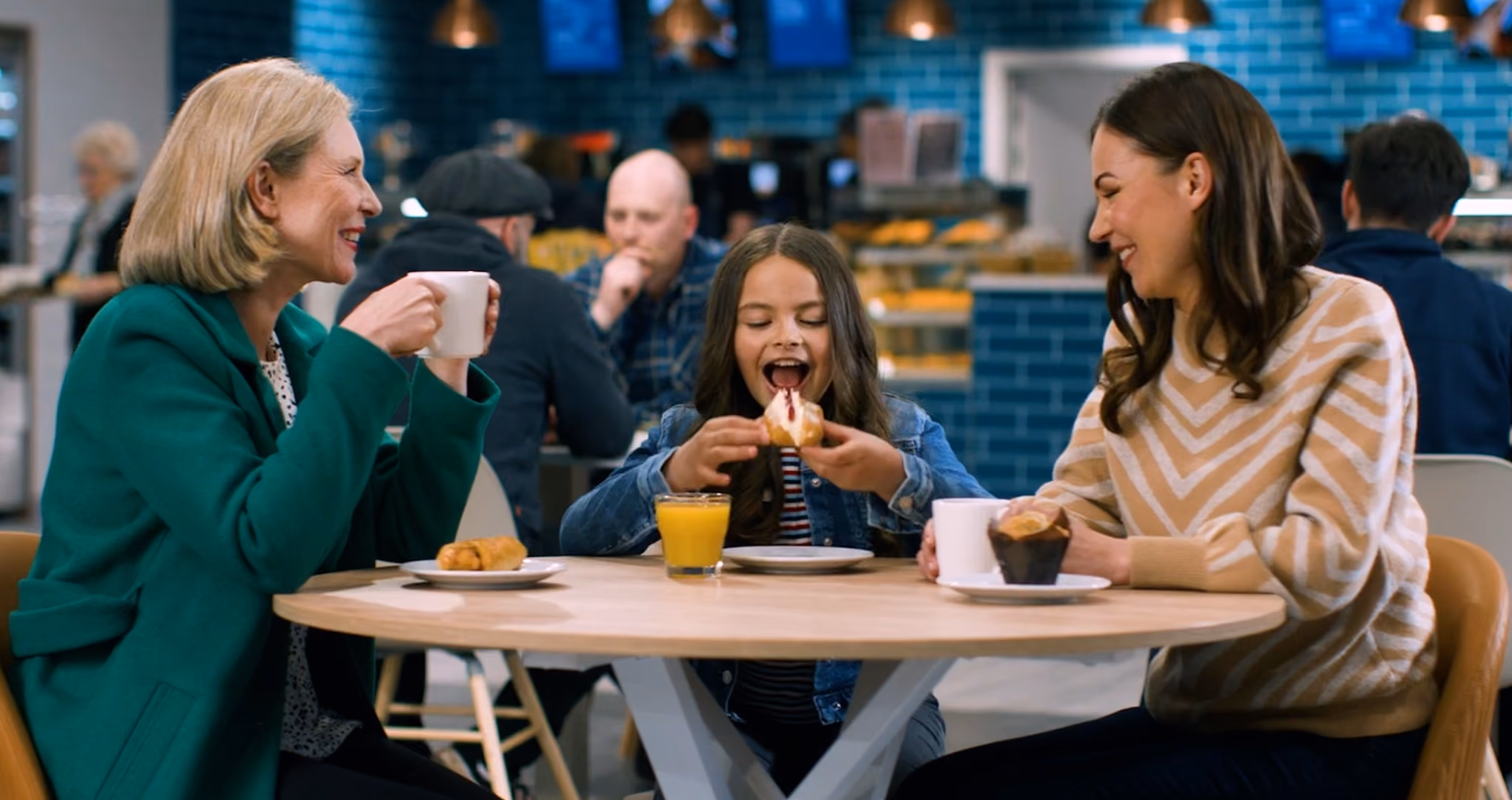 Three people sitting at a round table in a cafe; a girl in the middle is about to eat a sandwich, while two women on either side smile and hold cups, with pastries and a glass of juice on the table.