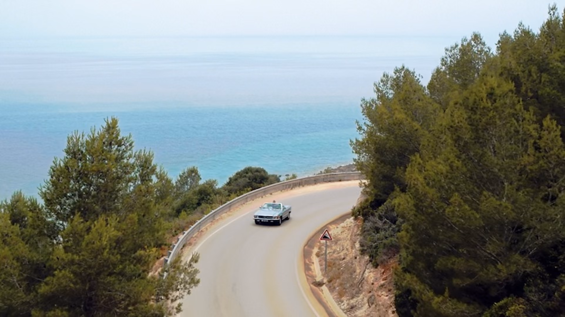 Vintage car driving on a winding coastal road surrounded by green trees with a view of the blue sea in the background.