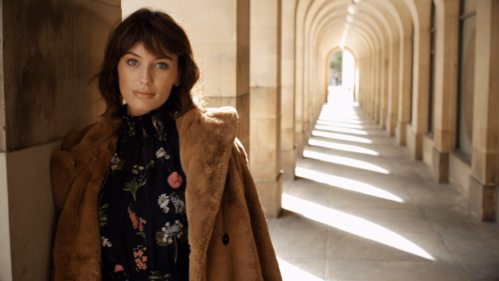 Woman with blue eyes and dark hair wearing a brown fur coat over a black floral dress, standing by a stone wall in a sunlit arched corridor.