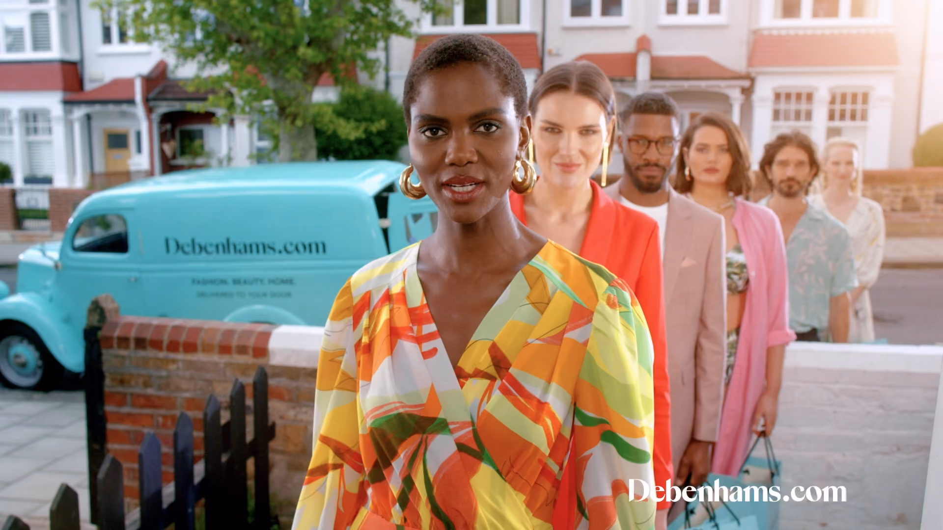 A diverse group of six fashionable adults standing in a line outdoors on a residential street, with a turquoise delivery van branded Debenhams.com in the background.