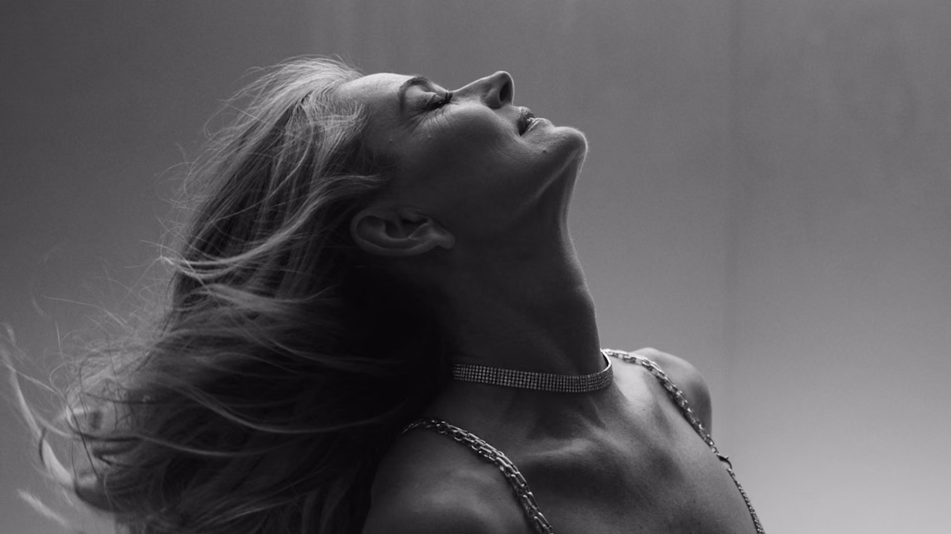 Black and white close-up of a model with windswept hair, head tilted back, wearing a delicate chain necklace, from a Karen Millen campaign