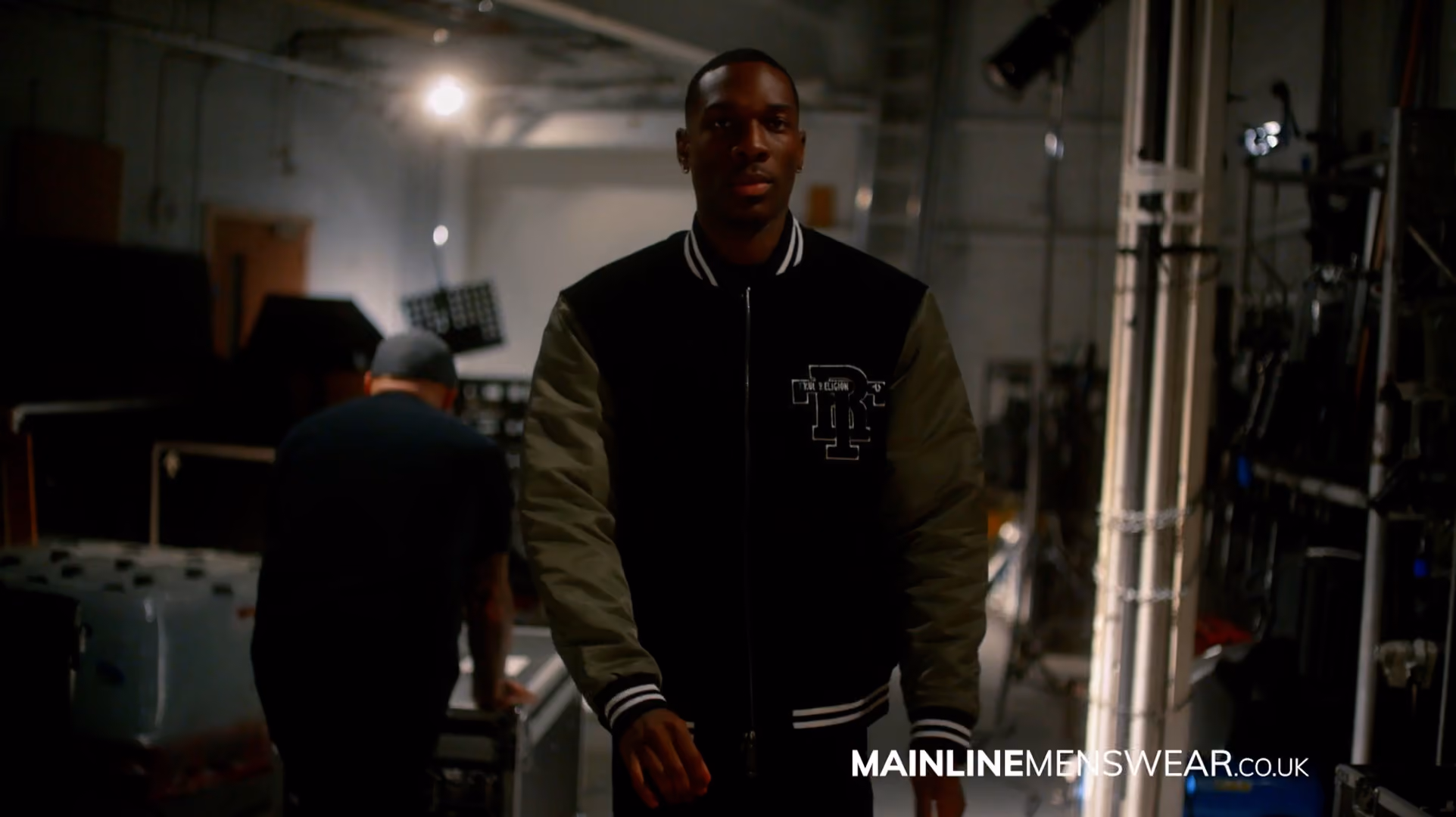 Man wearing a black and grey varsity jacket in an industrial warehouse setting, from a Mainline Menswear campaign