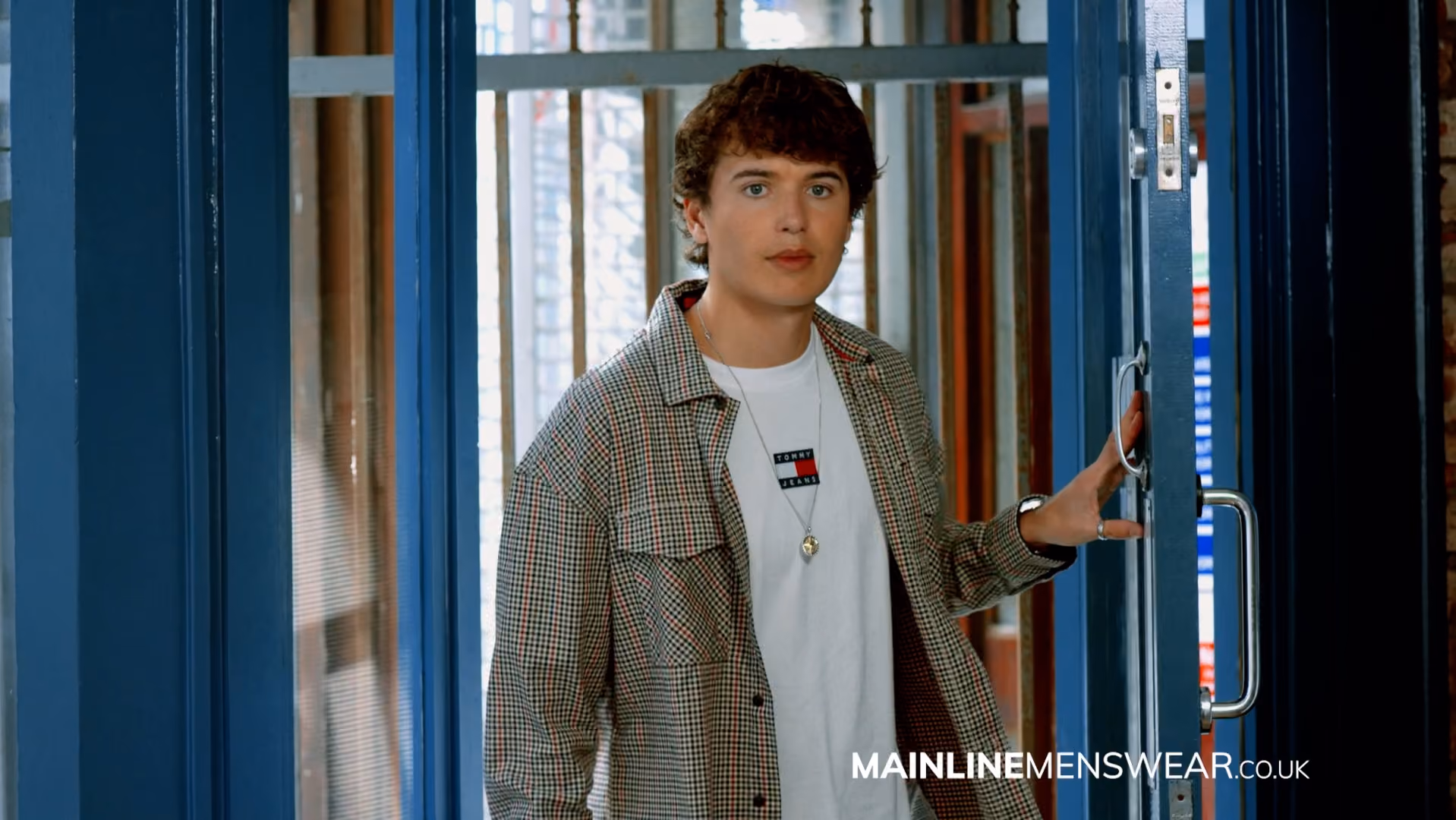 Teenage boy wearing a checked overshirt over a white t-shirt, standing in a doorway, from a Mainline Menswear campaign