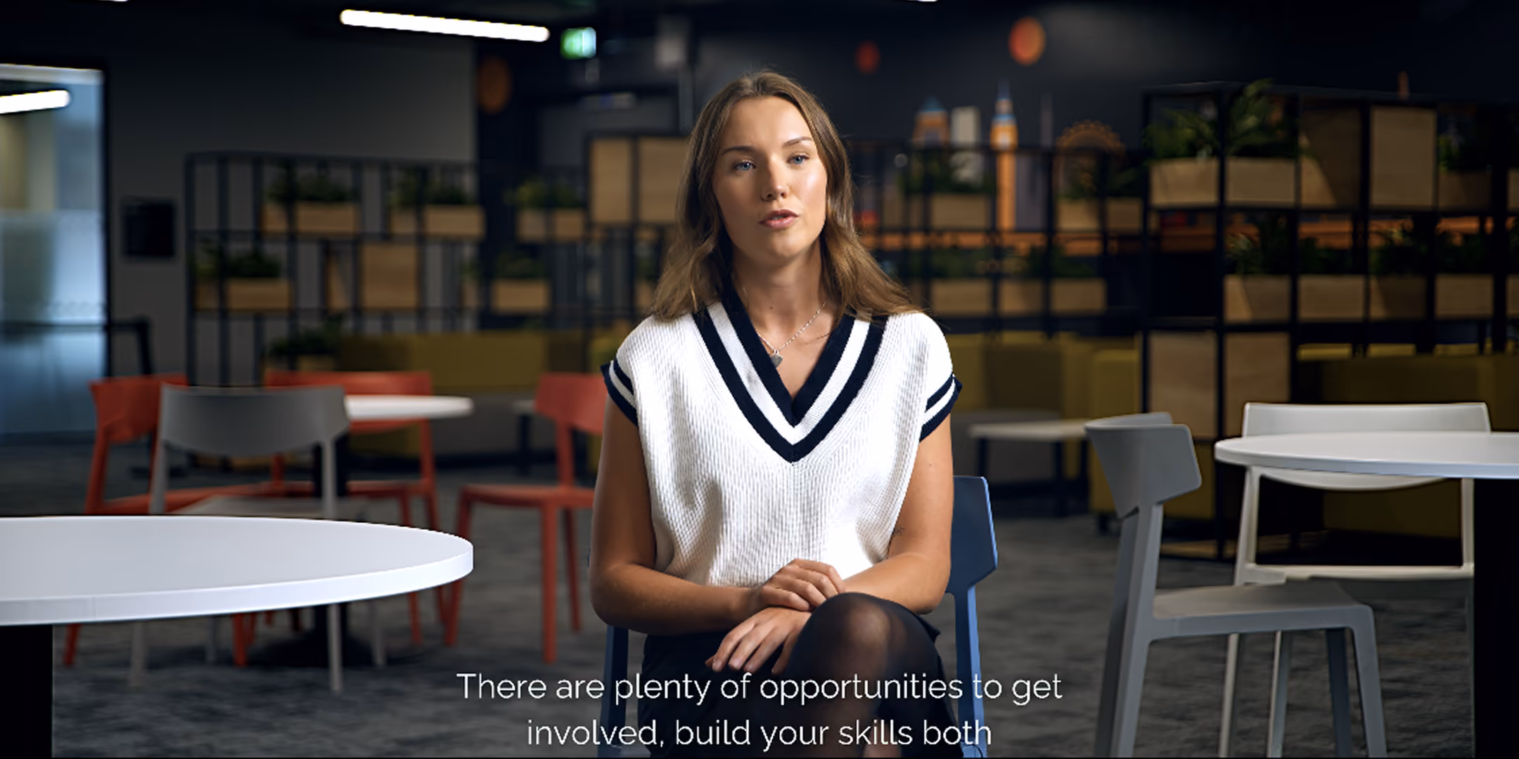 Young woman speaking to camera in a modern office breakout space, with on-screen subtitles
