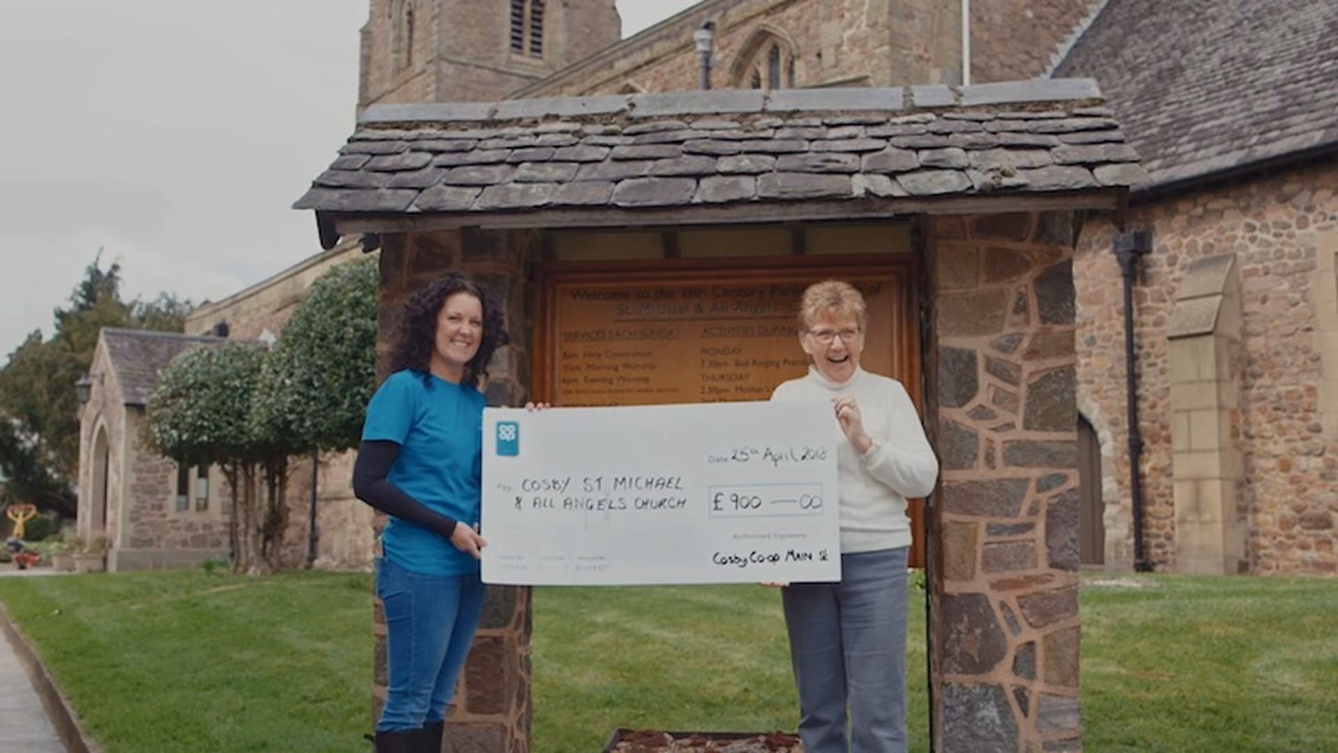 Two women holding a large charity cheque outside a church, marking a community fundraising presentation