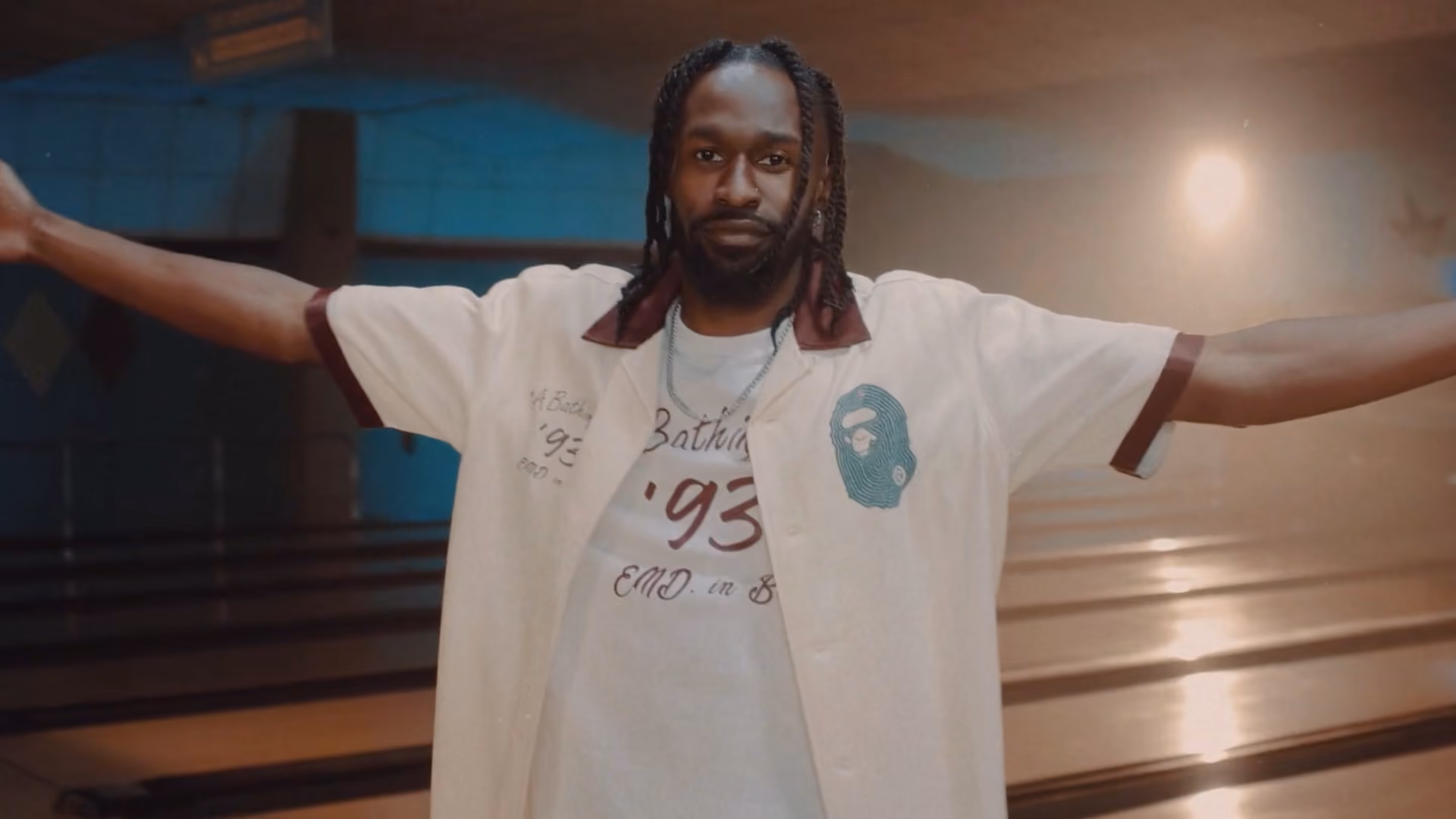 Man with dreadlocks wearing a white shirt with maroon trim and a graphic, standing with arms outstretched in a dimly lit bowling alley.