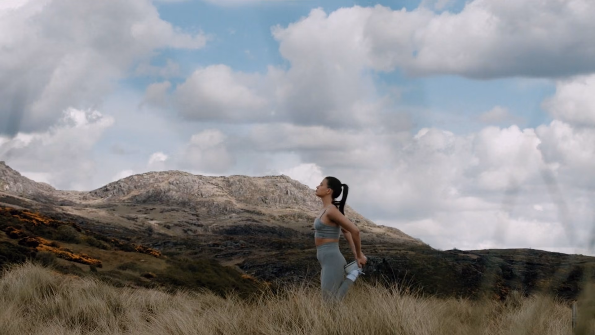 Woman in athletic clothes stretching her leg behind her and holding a water bottle, standing in tall grass with rocky hills and a partly cloudy sky in the background.