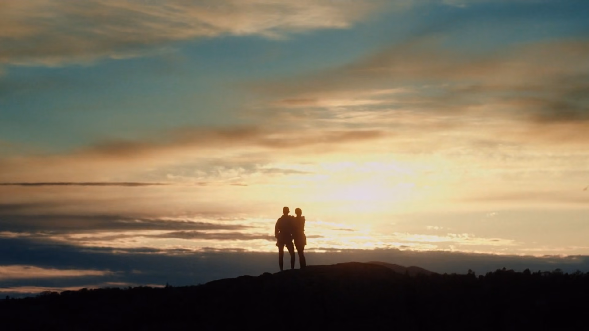 Silhouettes of two people standing on a hill against a sunset sky with scattered clouds and warm light.