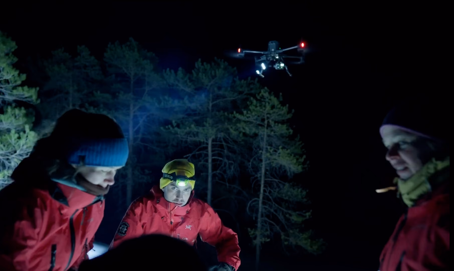 Three people wearing red jackets and winter hats gathered outdoors at night with a drone flying above them illuminating the scene and trees visible in the background.