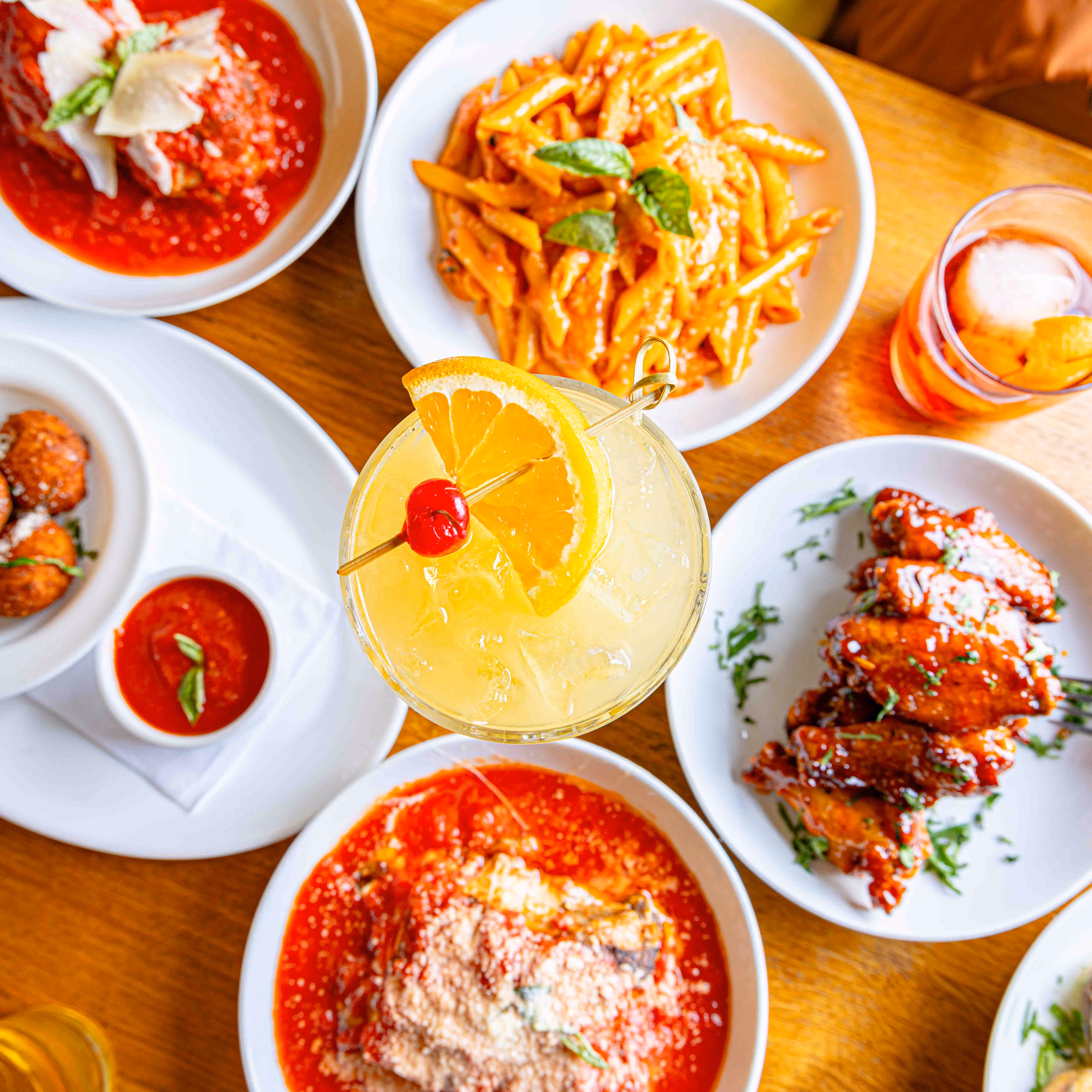 Italian plates on a wooden table at Adrienne's Pizzabar, including baked pasta, stuffed mushrooms, and crostini, with a glass of white wine in front of a dark leather booth