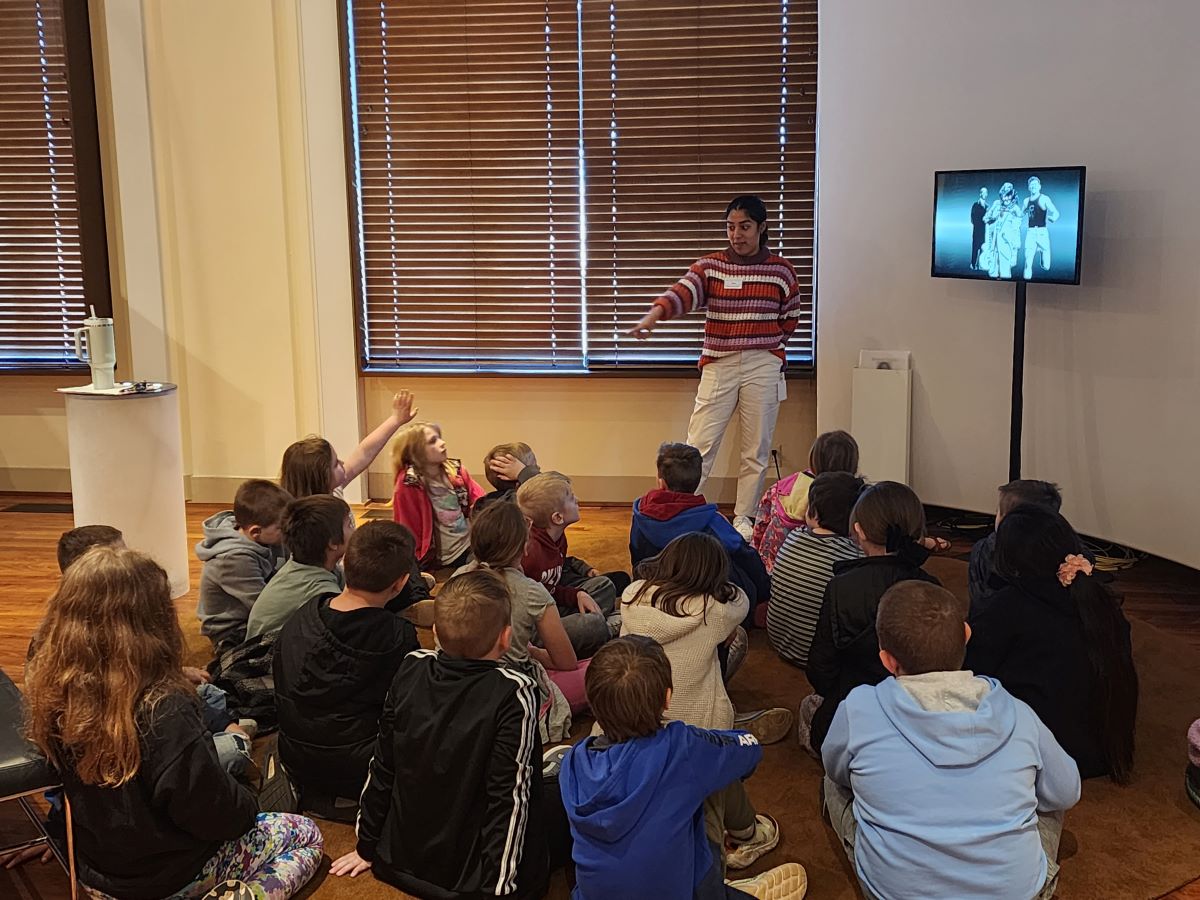 Teacher standing and pointing while a group of children sit on the floor in a classroom with a monitor displaying a black and white image.