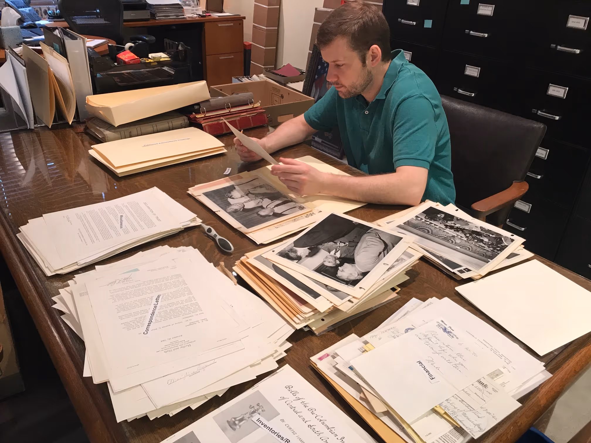 Man in green shirt sitting at a desk reviewing historical documents and black-and-white photographs.