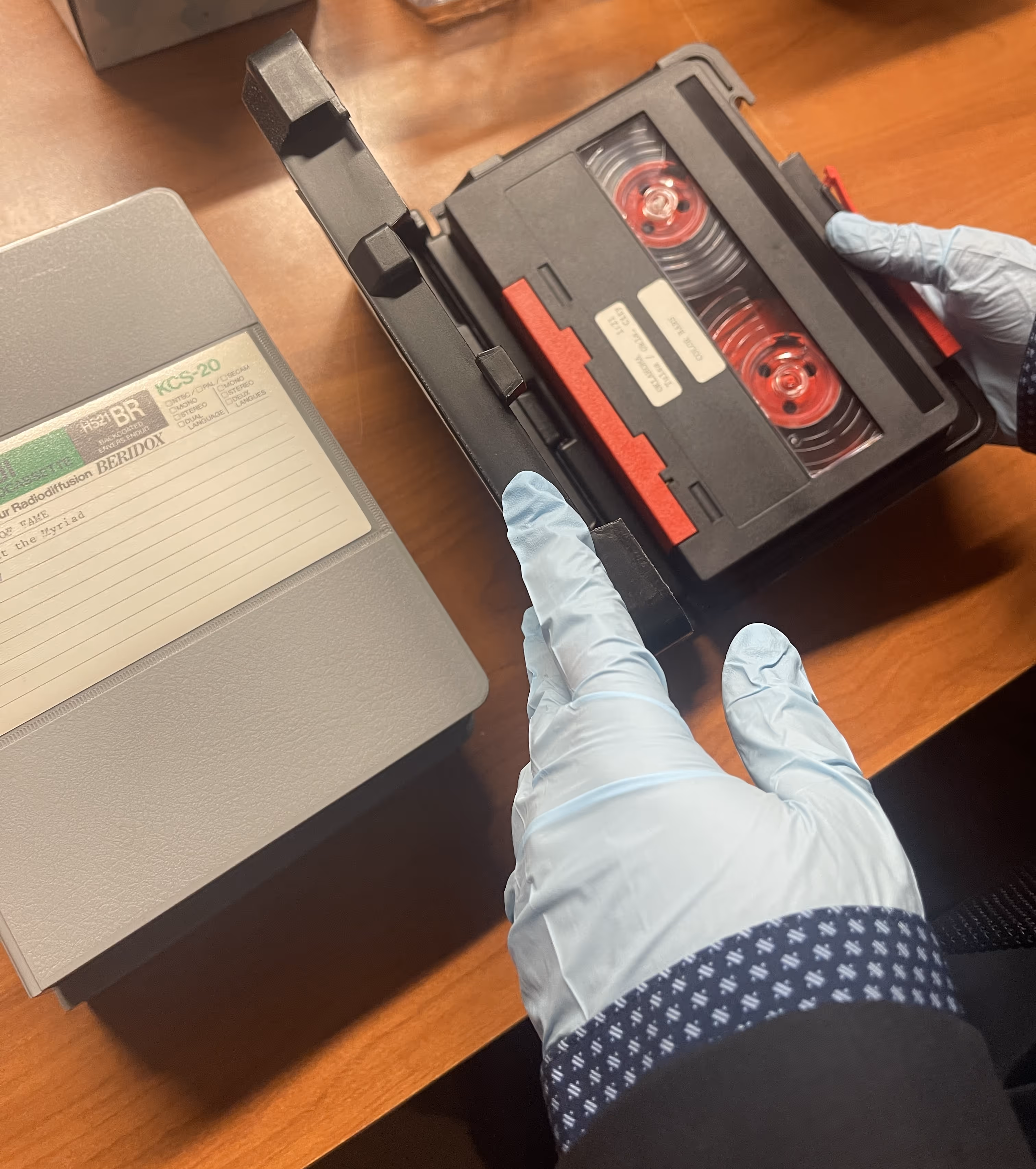 Hands in blue gloves holding an open black VHS tape case with a tape inside on a wooden surface.