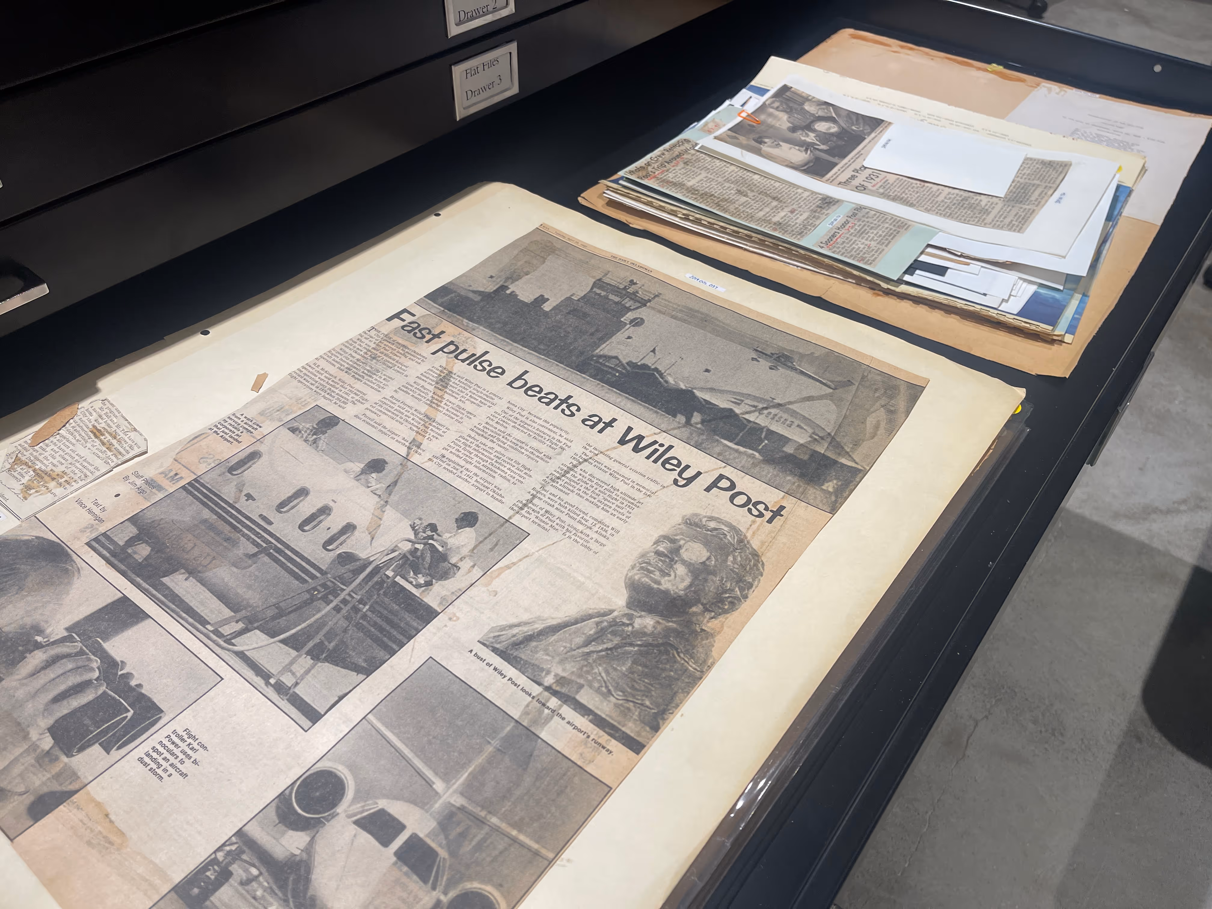 Archive drawer open displaying old newspaper page titled 'Fast pulse beats at Wiley Post' with aircraft images and a bust statue photo.