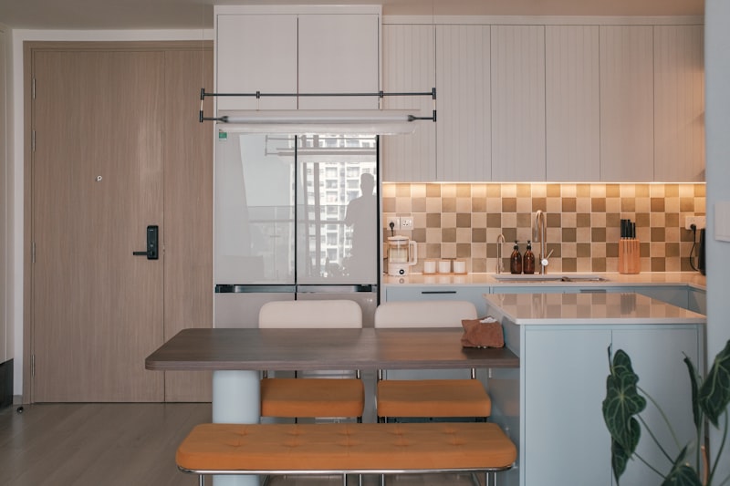 A bright modern apartment kitchen and dining area with a light-wood door and tiled backsplash