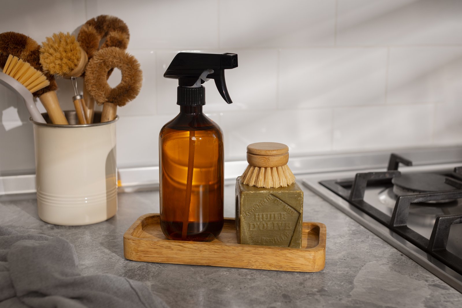 An amber glass spray bottle, wooden scrubbing brushes and a block of soap on a grey kitchen worktop