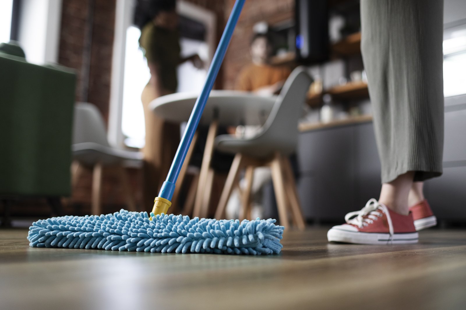 A blue microfibre flat mop being pushed across a wooden floor in a bright dining area