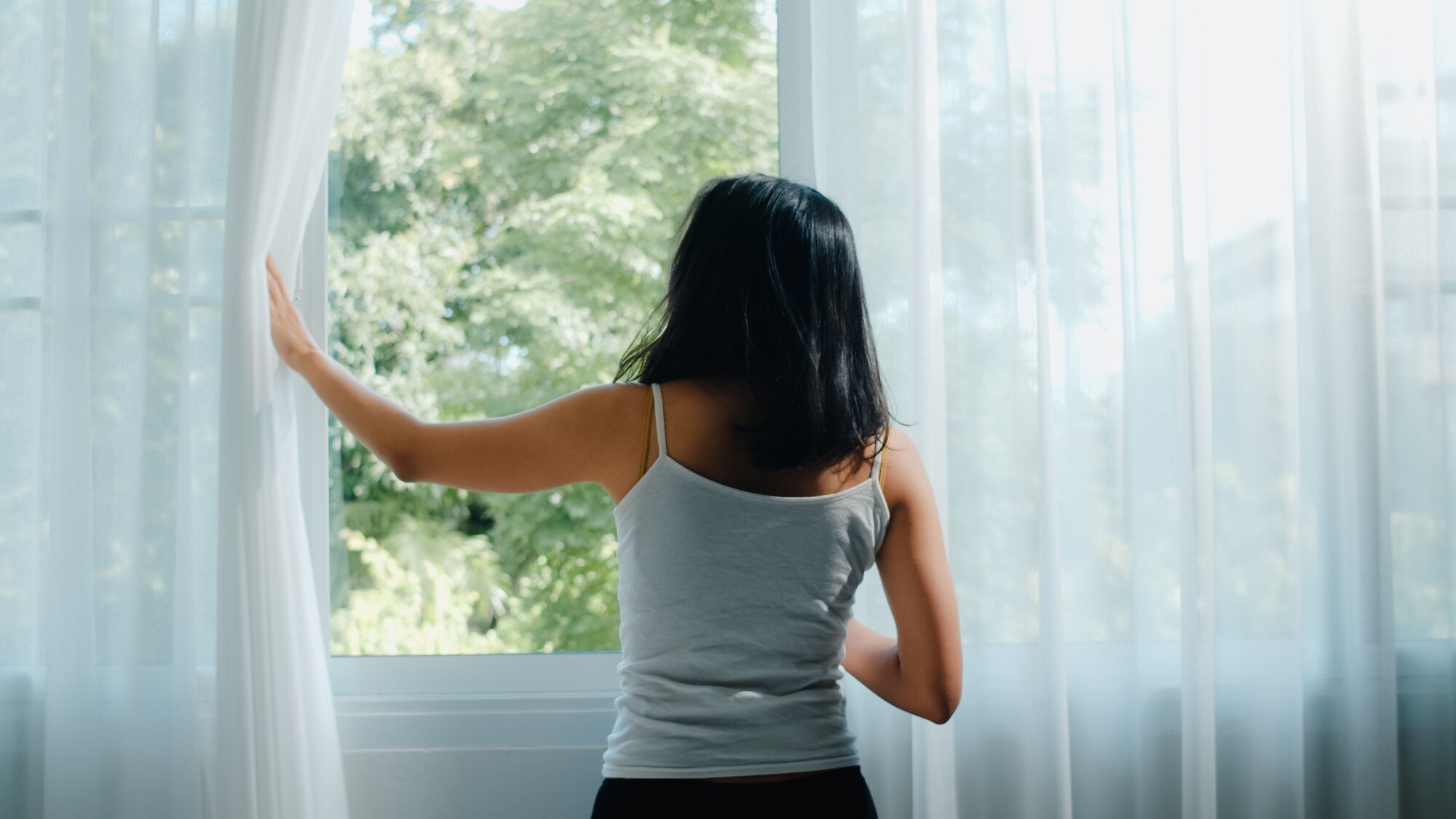 A woman drawing back sheer curtains to open a window and air out a room after time away