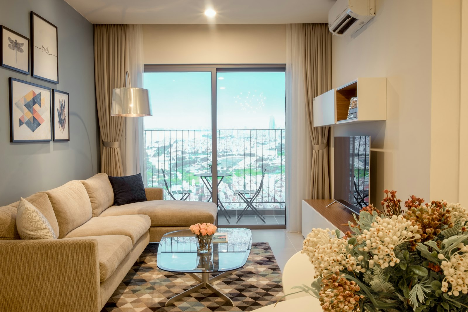 Bright high-rise flat living room with a beige fabric sofa, patterned rug, balcony doors and a wall-mounted aircon unit