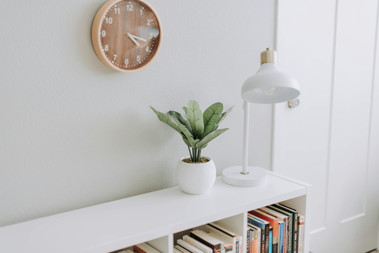 A white wall with a wooden clock, a desk lamp and a small potted plant above a low shelf of books