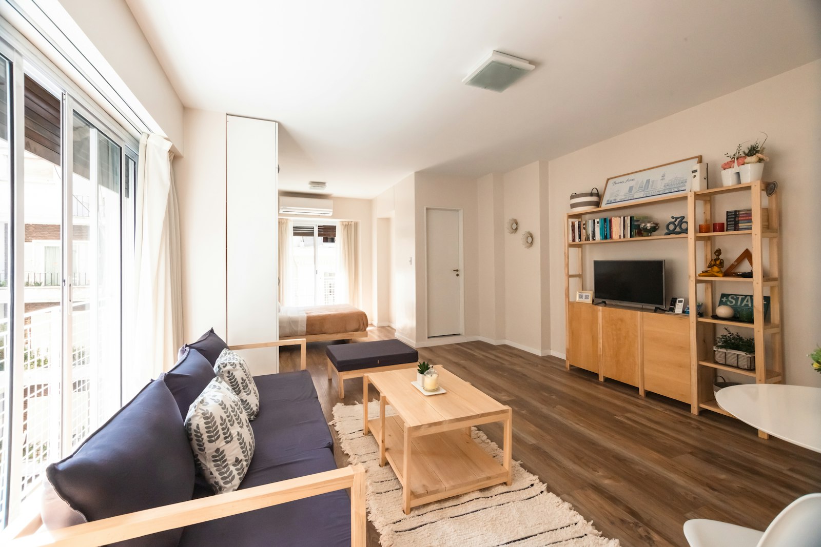 A bright apartment living room with a wooden floor, low sofa, woven rugs and a sliding door to the balcony