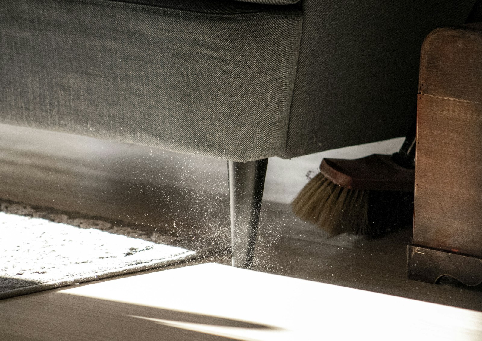 A cloud of dust being swept out from underneath a fabric sofa with a soft brush