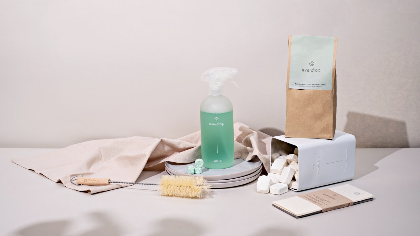 Cleaning supplies on a pale table — a bottle of green liquid soap, a folded cloth and paper packaging